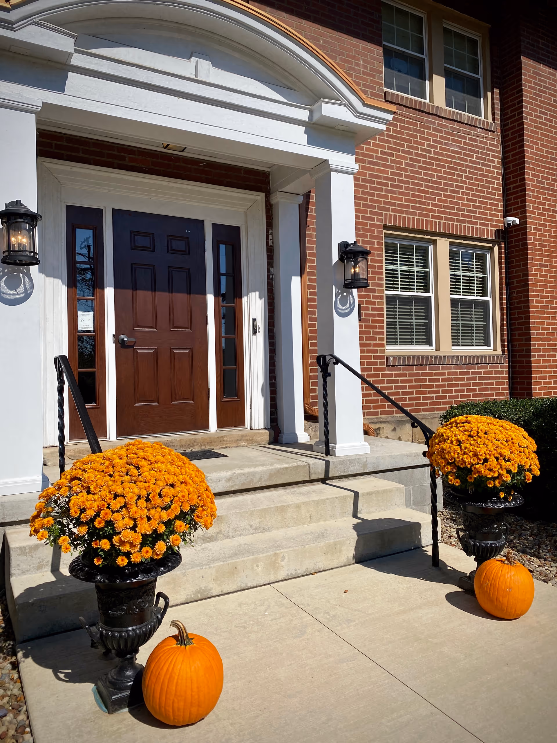 Entrance to a brick building with a dark brown door framed by white columns and a decorative arch. Two large pots of orange flowers and two pumpkins are placed symmetrically on either side of the concrete steps leading up to the door. Black metal handrails flank the steps, and two black lantern-style wall lights are mounted on the white columns.
