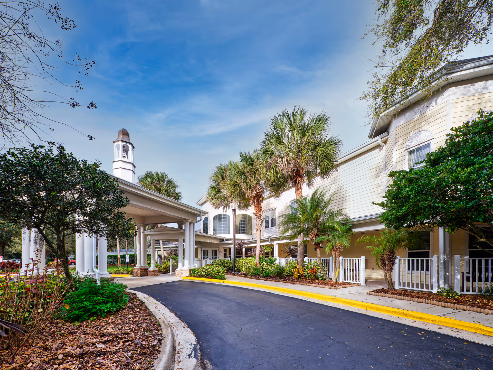 Exterior view of Colliers Assisted Living at Port Orange showing a covered entrance with white columns, palm trees, and landscaping along a curved driveway under a blue sky.