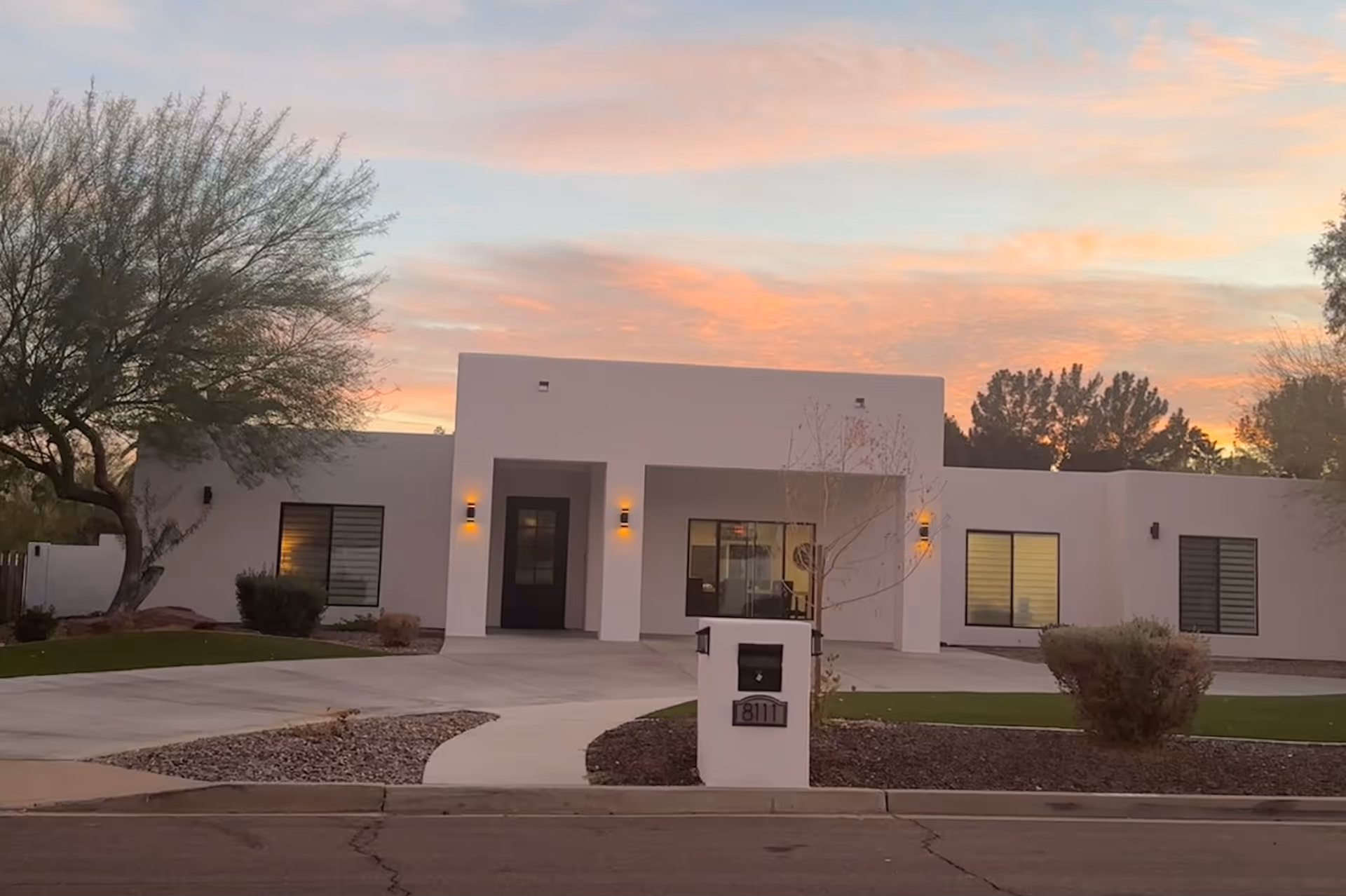 Front exterior view of a modern, single-story building with a flat roof and white walls, featuring a central entrance with two wall-mounted lights on either side. The building is surrounded by a landscaped yard with green grass, bushes, and a tree on the left side. The sky is colorful with a sunset glow.
