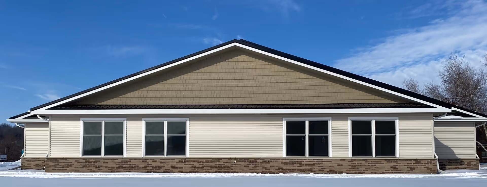 Single-story beige assisted-living building façade with multiple large windows, brick trim, and snow in the foreground under a blue sky.