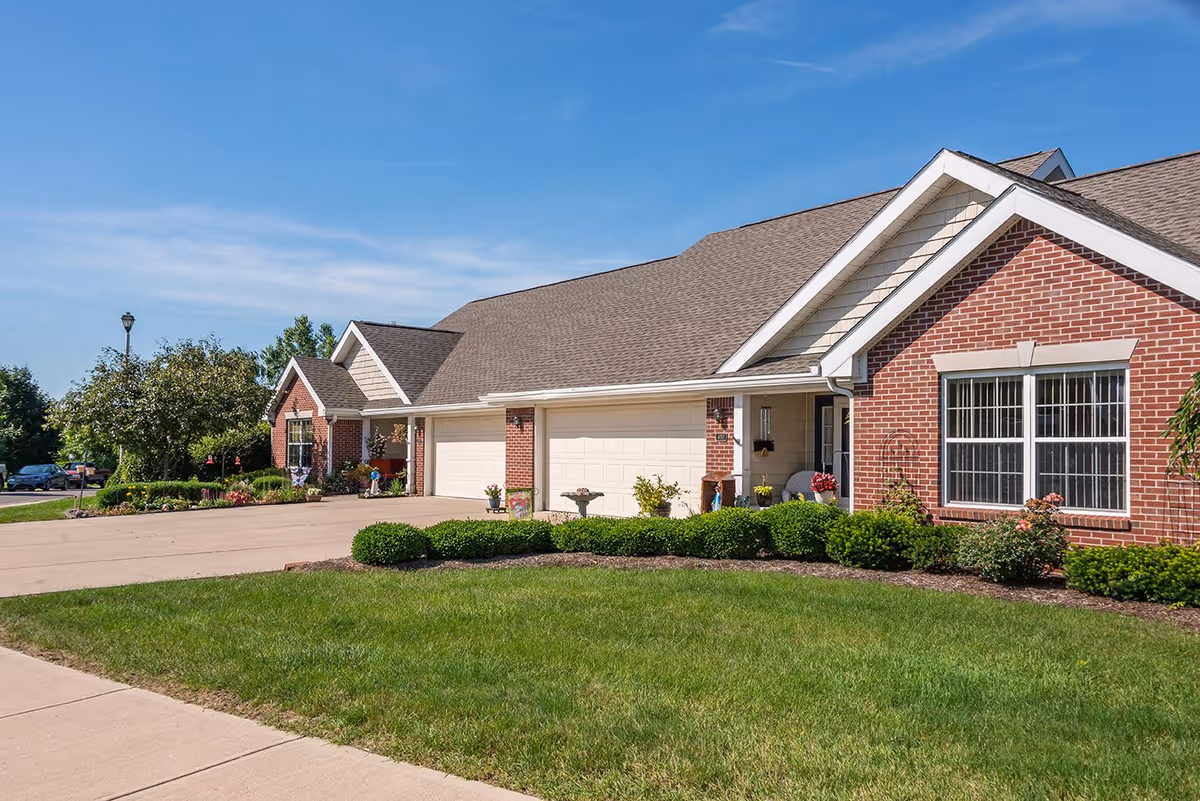 Exterior view of a single-story senior living facility with brick walls, multiple garages, well-maintained green lawn, shrubs, and a clear blue sky.