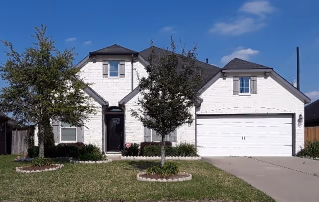Front view of a two-story white brick suburban house with a two-car garage, driveway and small front lawn with trees.