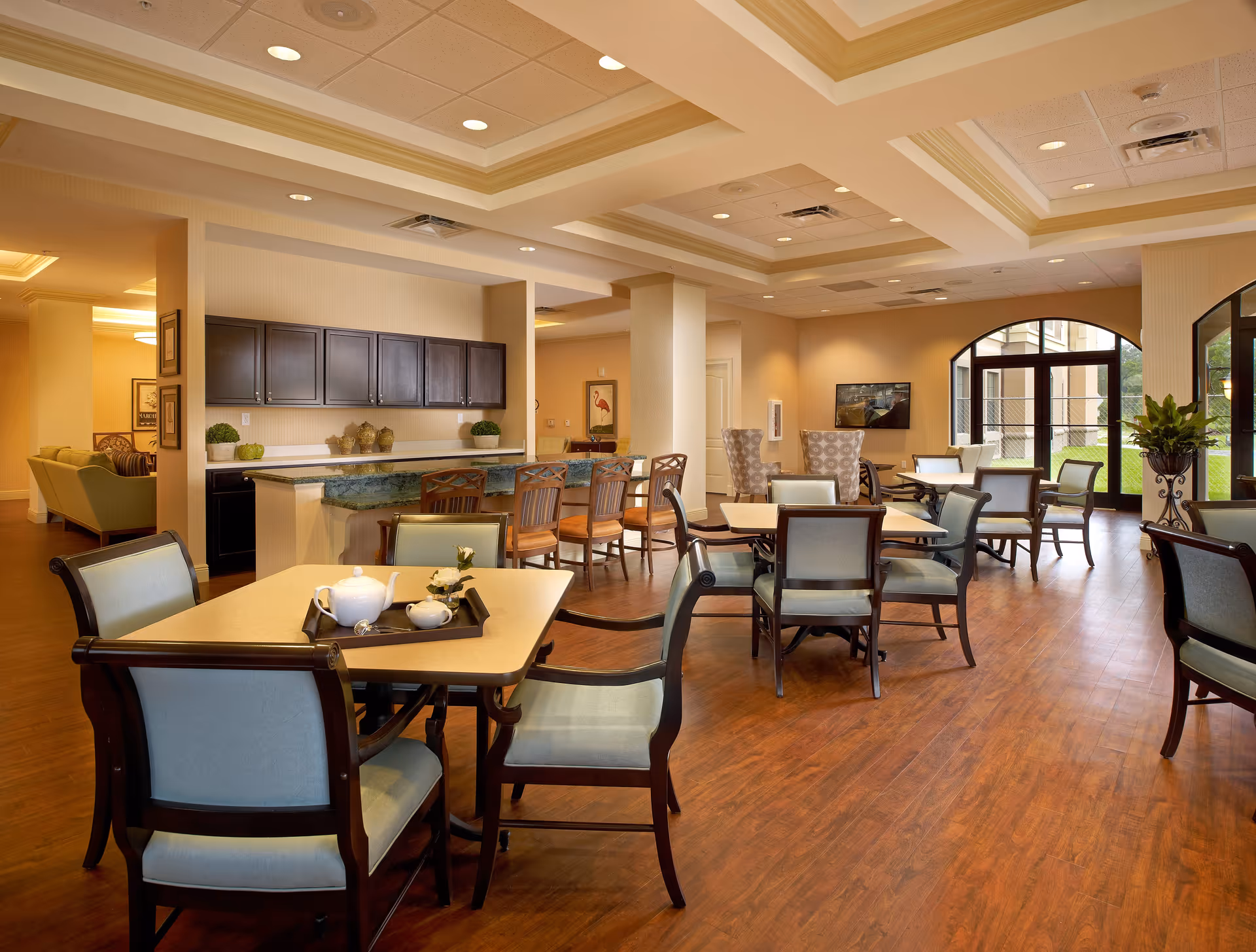 A spacious and well-lit common area in a senior living facility featuring multiple tables and chairs arranged for dining or socializing. The room has wooden flooring, a ceiling with recessed lighting, and a counter with bar stools. Large windows and glass doors allow natural light to enter, and there are plants and framed artwork decorating the space.