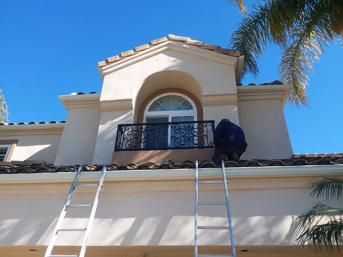 Facade of a stucco two-story house with an arched window and small wrought-iron balcony, a worker kneeling on the tiled roof and two ladders leaning against the eaves.