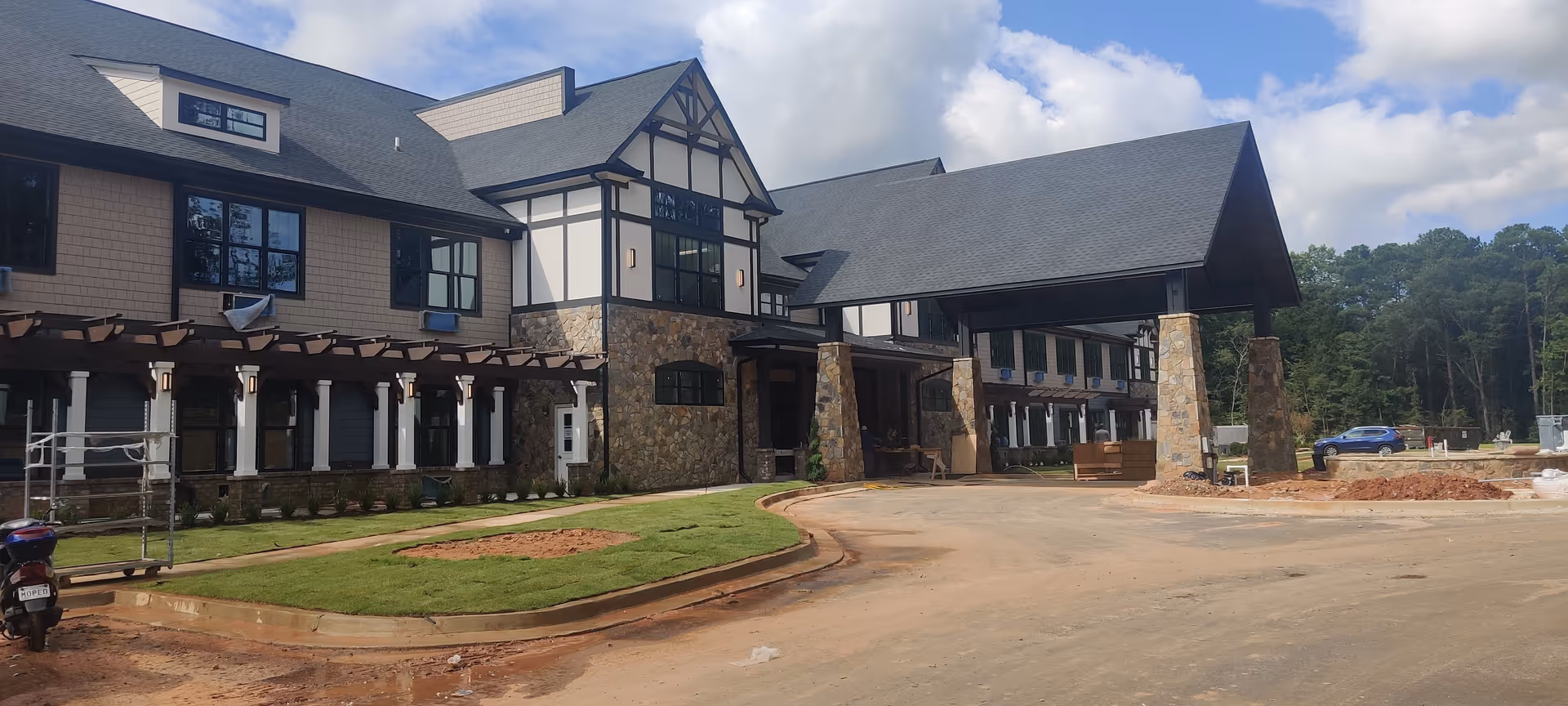 Exterior view of a large senior living facility building with stone and wood accents, a covered entrance supported by stone pillars, newly laid grass, and a dirt driveway under a partly cloudy sky.