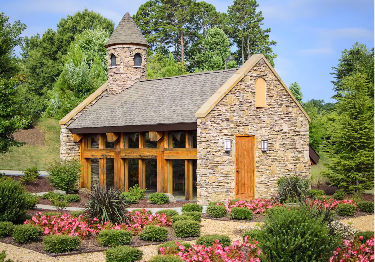 Stone cottage-style building with a wooden door and large windows set amid landscaped gardens and flowering shrubs.