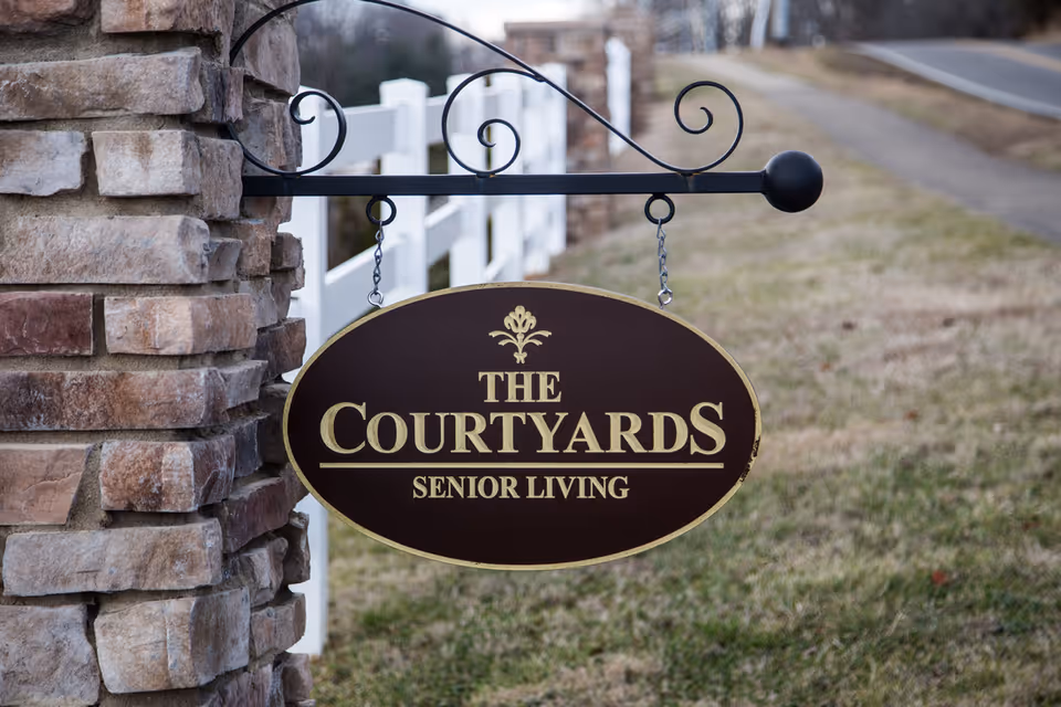 A brown oval sign with gold lettering that reads 'THE COURTYARDS SENIOR LIVING' hanging from a decorative black metal bracket attached to a stone pillar, with a white fence and a road in the background.