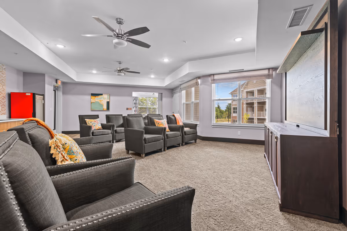 A spacious senior living lounge with black armchairs, ceiling fans, and a large window.