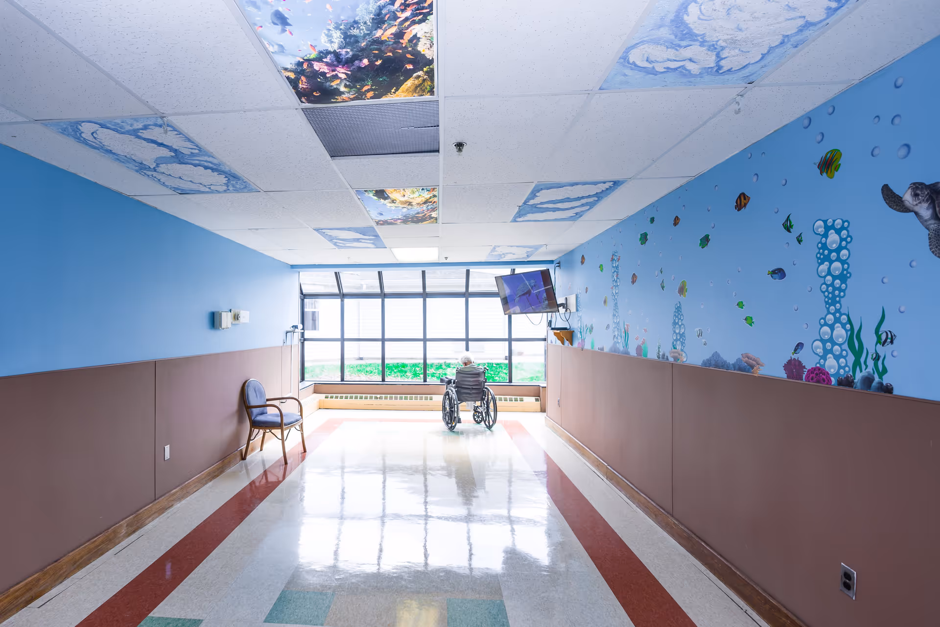 A bright hallway in a senior facility with an ocean-themed mural, decorative ceiling panels, and a wheelchair positioned by a large window.