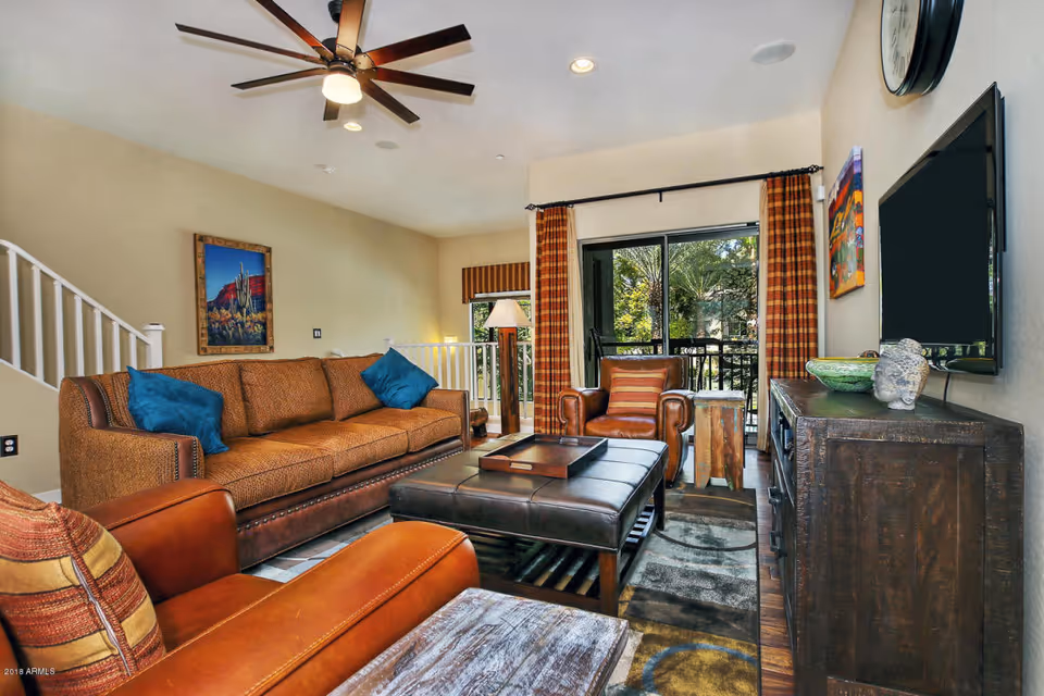 A cozy living room with a brown sofa adorned with blue pillows, two brown leather armchairs with striped cushions, a large dark brown leather ottoman coffee table, a wooden side table, and a dark wooden TV stand with a flat-screen TV mounted on the wall. The room features a ceiling fan with lights, a large window with plaid curtains overlooking greenery, and a framed picture on the wall.