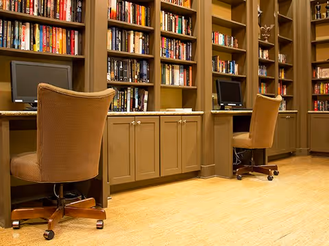 A quiet library or study area with built-in bookshelves filled with books, two computer workstations with monitors, and two brown upholstered office chairs on wheels. The room has wooden flooring and warm lighting.
