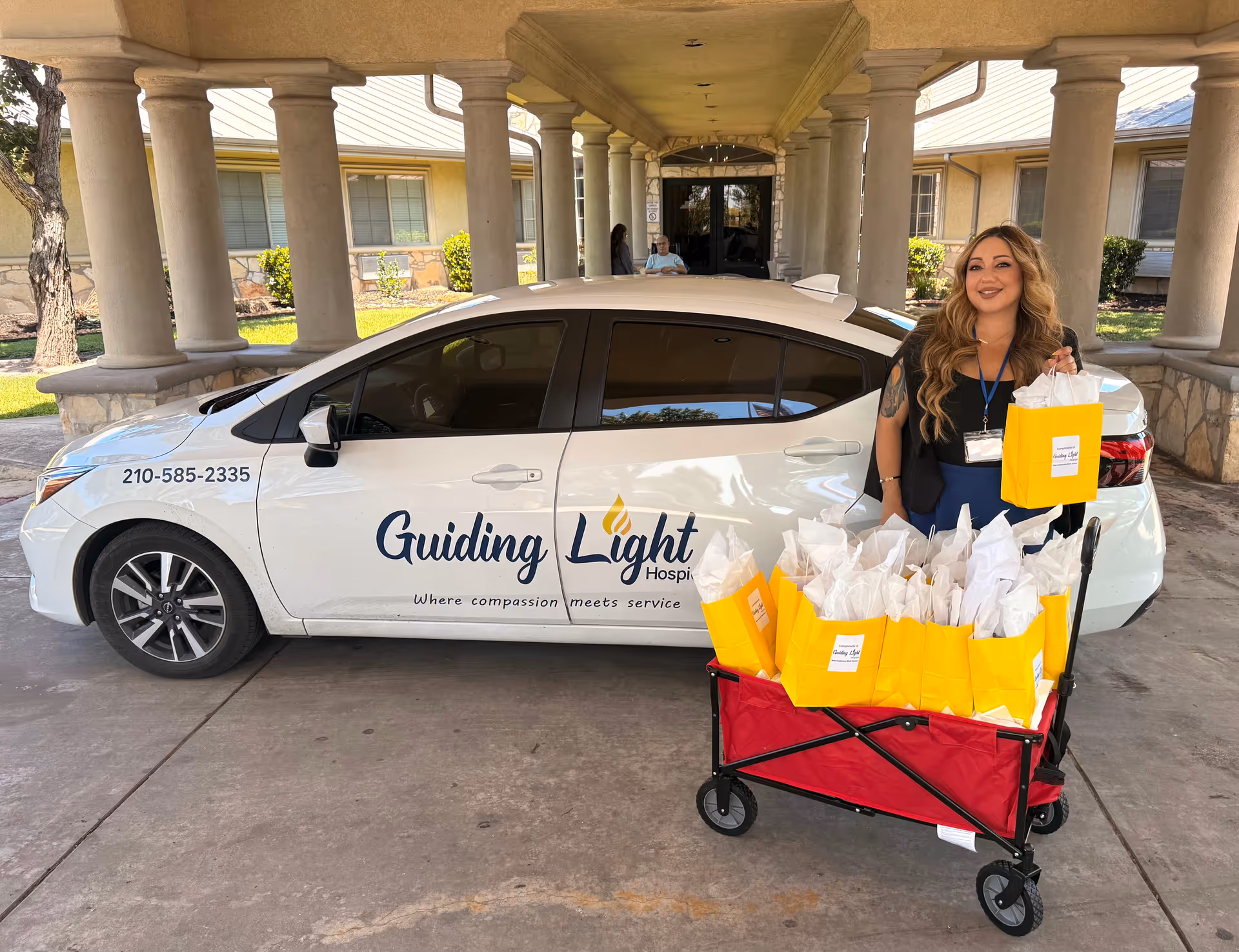 Staff member stands beside a white Guiding Light Hospice car with a wagon of gift bags.