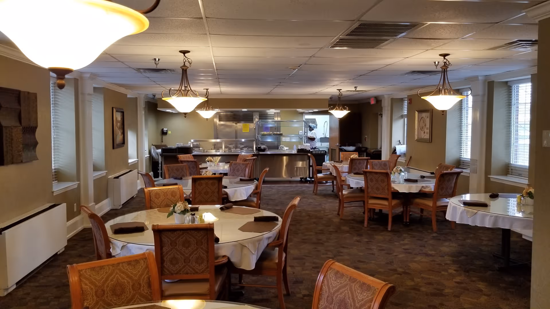 Dining room with round tables covered in white tablecloths and upholstered chairs facing a serving area and kitchen.