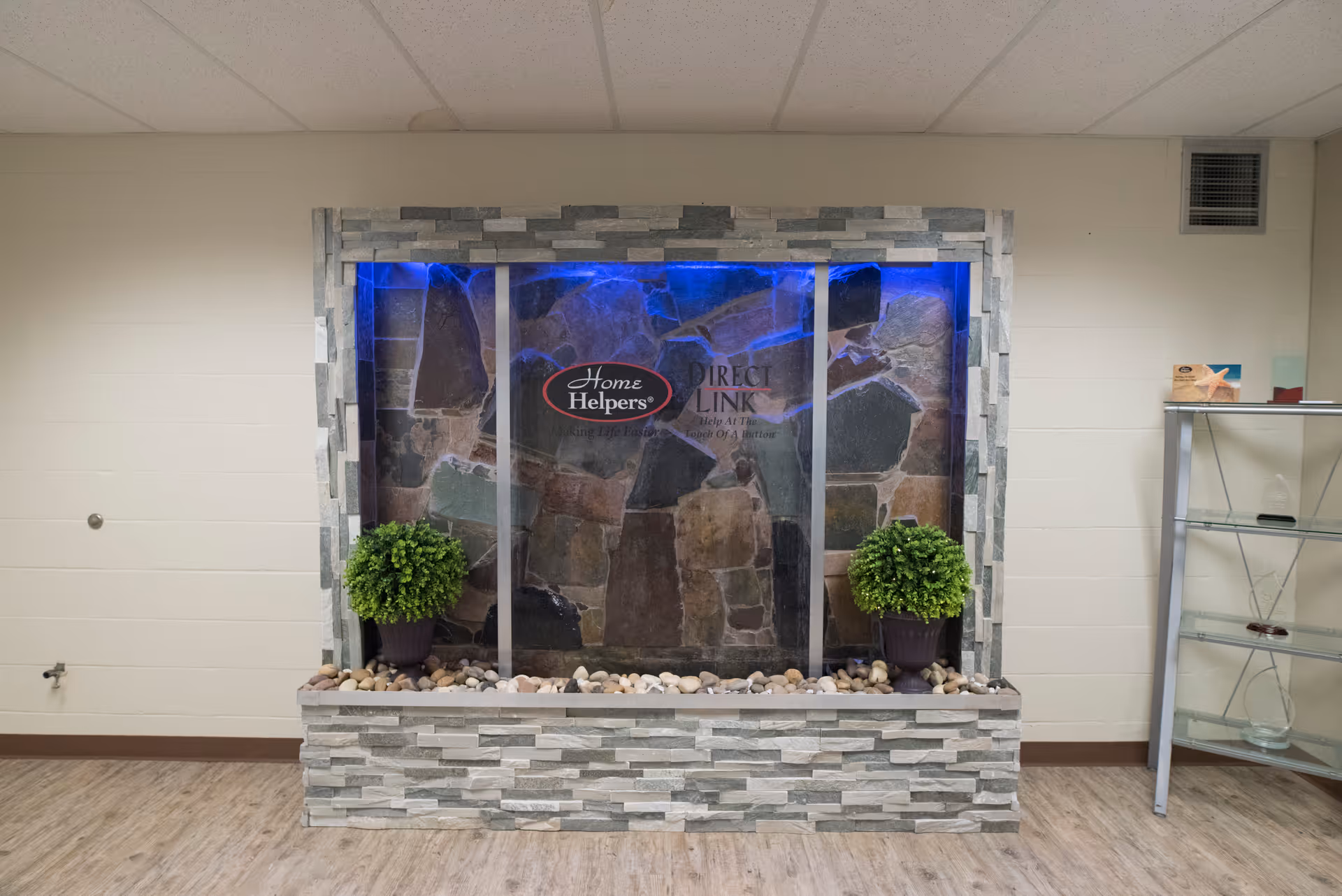 Stone water feature with glass panels, blue light, and two potted plants in a hallway.