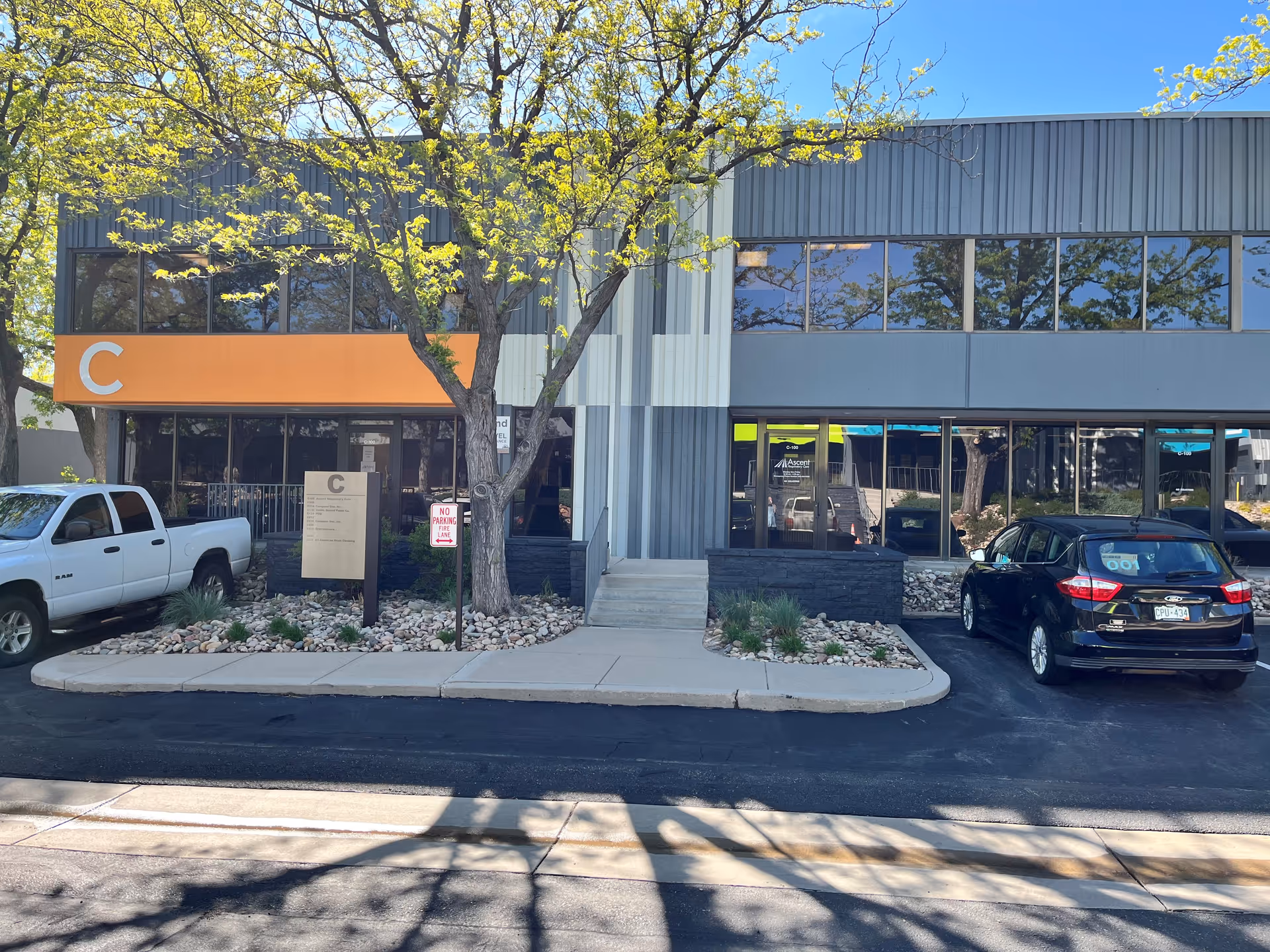 Exterior view of Ascent Respiratory Care building with glass entry, orange C sign, and parked vehicles.