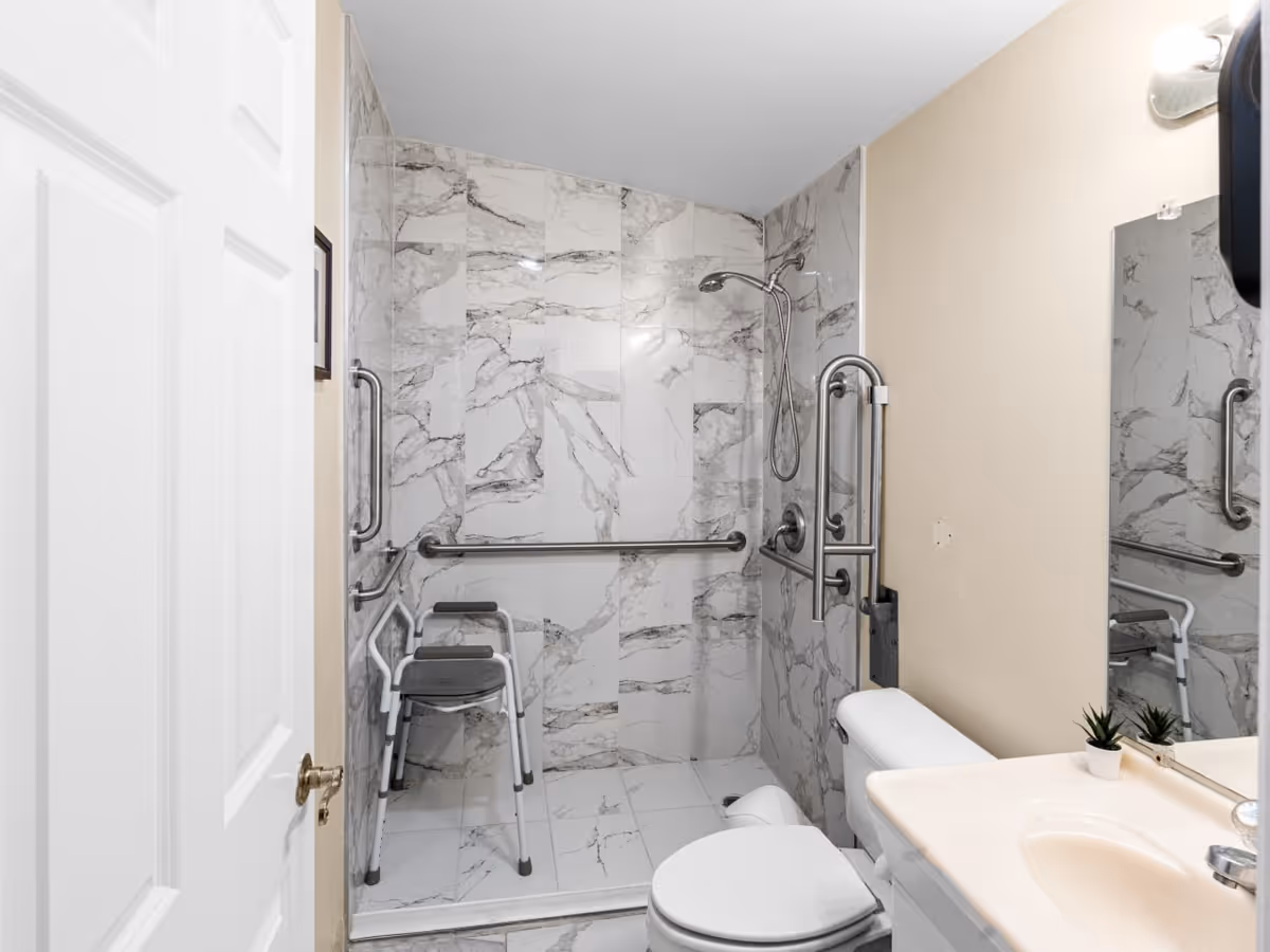 Accessible marble-tiled bathroom with grab bars, a seated shower, toilet, and sink.