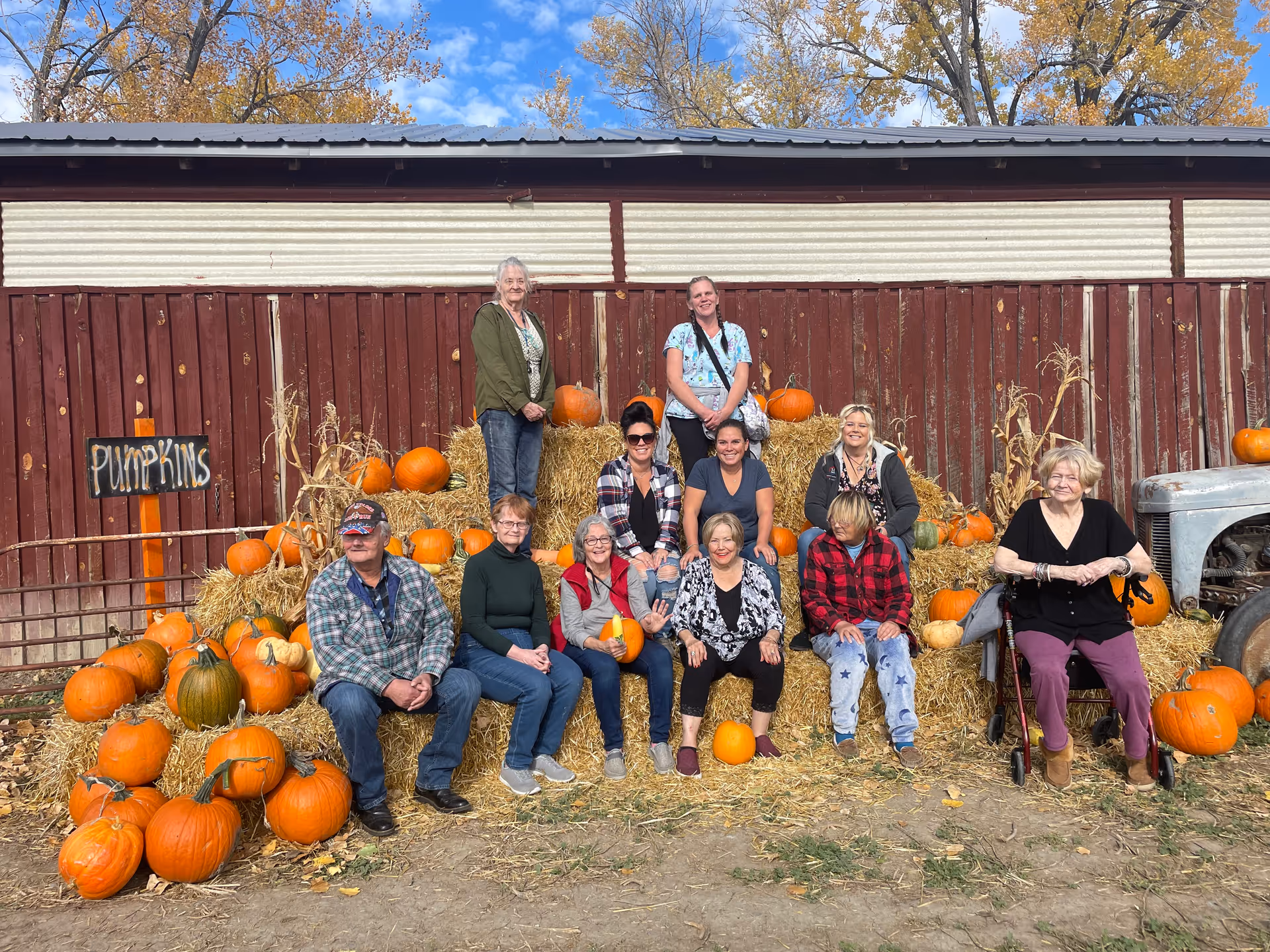 A group of eleven people posing outdoors in front of a rustic red barn wall decorated with hay bales and numerous pumpkins. The group includes men and women of various ages, some sitting on the hay bales and others standing behind. There is a sign that says 'PUMPKINS' on the left side, and a vintage tractor partially visible on the right. The scene is set on a sunny day with blue sky and autumn trees in the background.