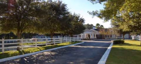 Sunlit driveway to Brookdale Orange Park entrance with white fencing and mature trees.