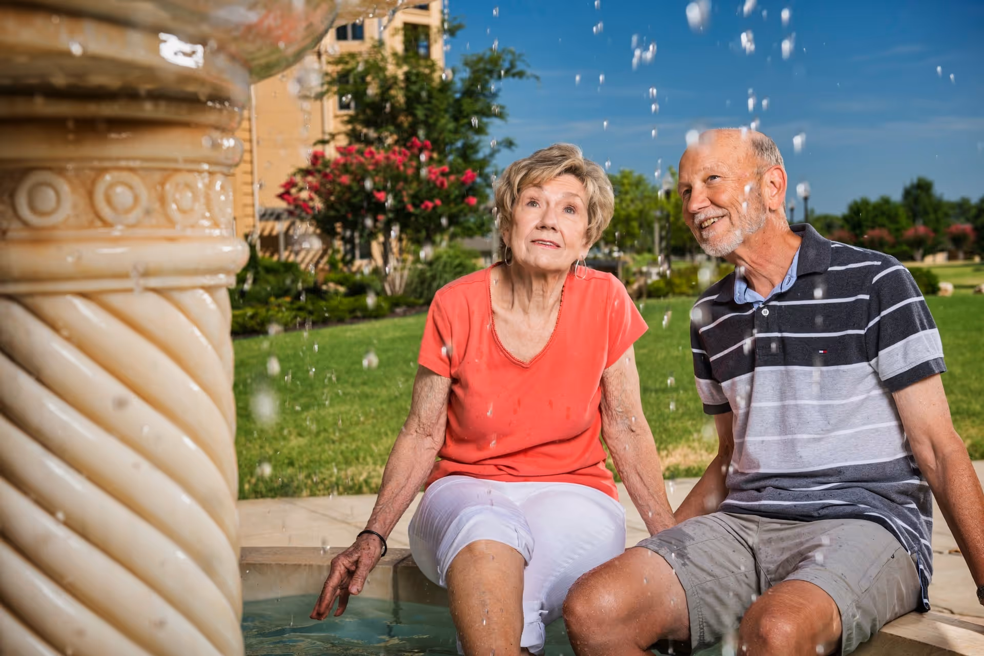 An elderly woman in a red shirt and white pants and an elderly man in a striped polo shirt and shorts sit by a water fountain outdoors on a sunny day, with green grass, trees, and a building in the background.