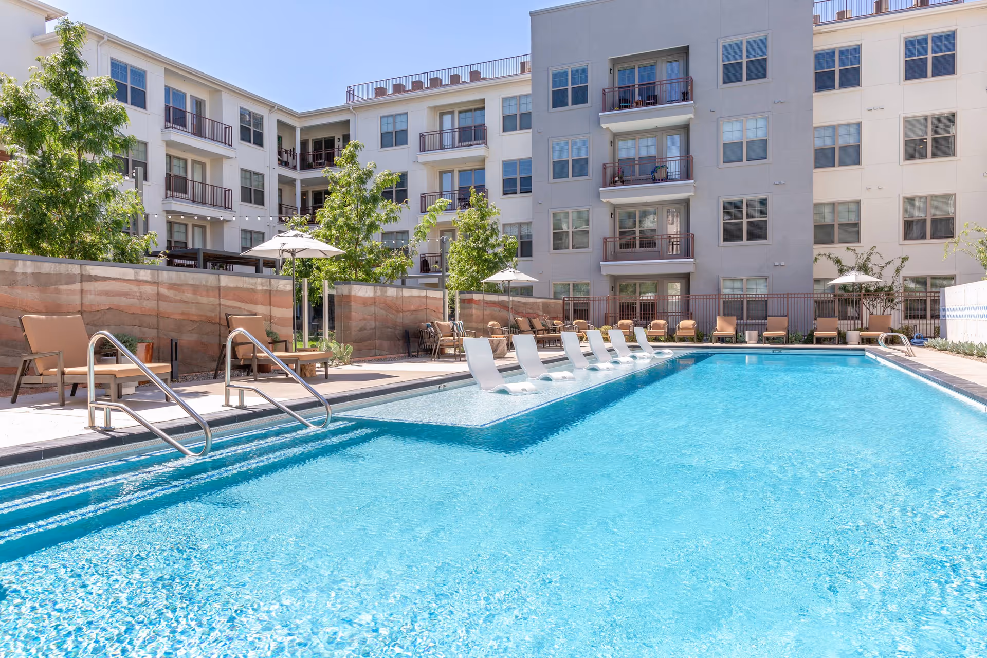 Outdoor swimming pool with in-pool lounge chairs, deck chairs and umbrellas in the courtyard of a multi-story residential building.