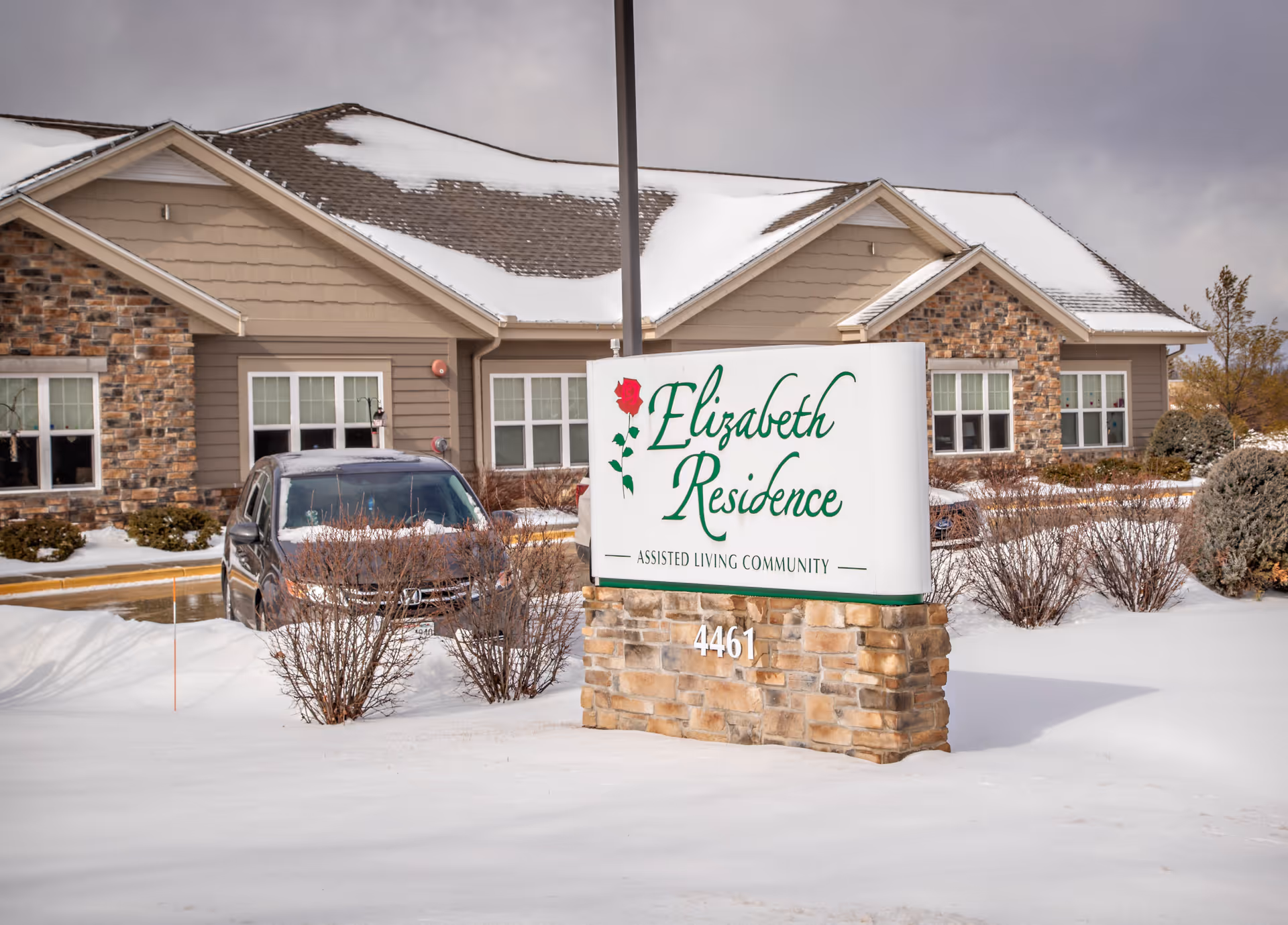 Exterior view of Elizabeth Residence assisted living community building in winter with snow on the ground and roof. A sign in front displays the facility name and address 4461. A car is parked near the building.