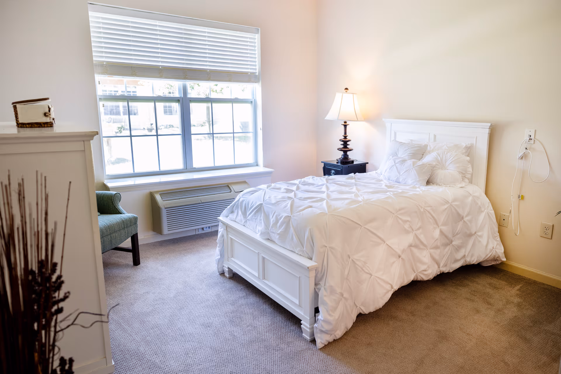 Bright bedroom with white bed, nightstand, lamp, and window seating area.