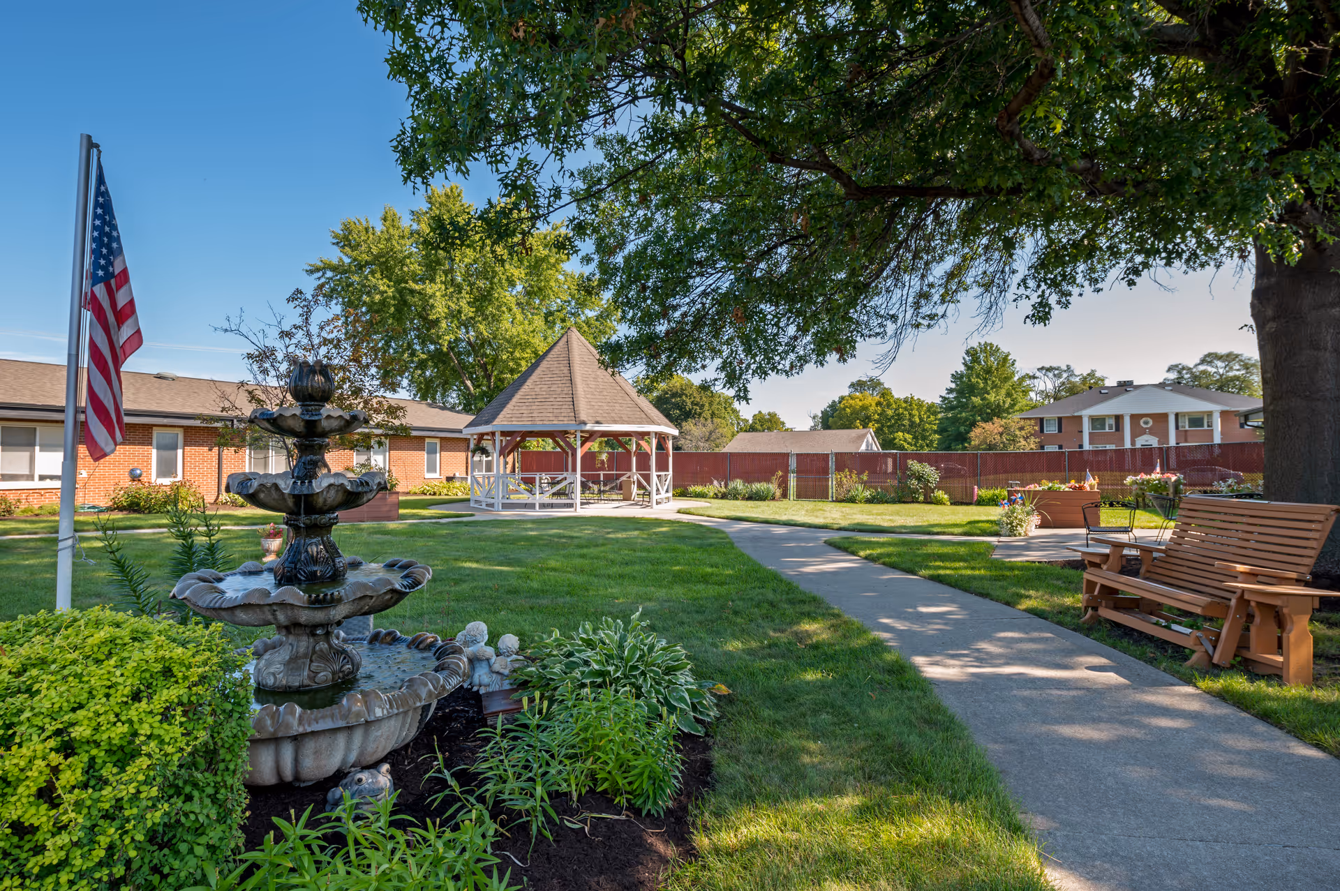 A well-kept outdoor courtyard with fountain, gazebo, benches, flag, and walking path.