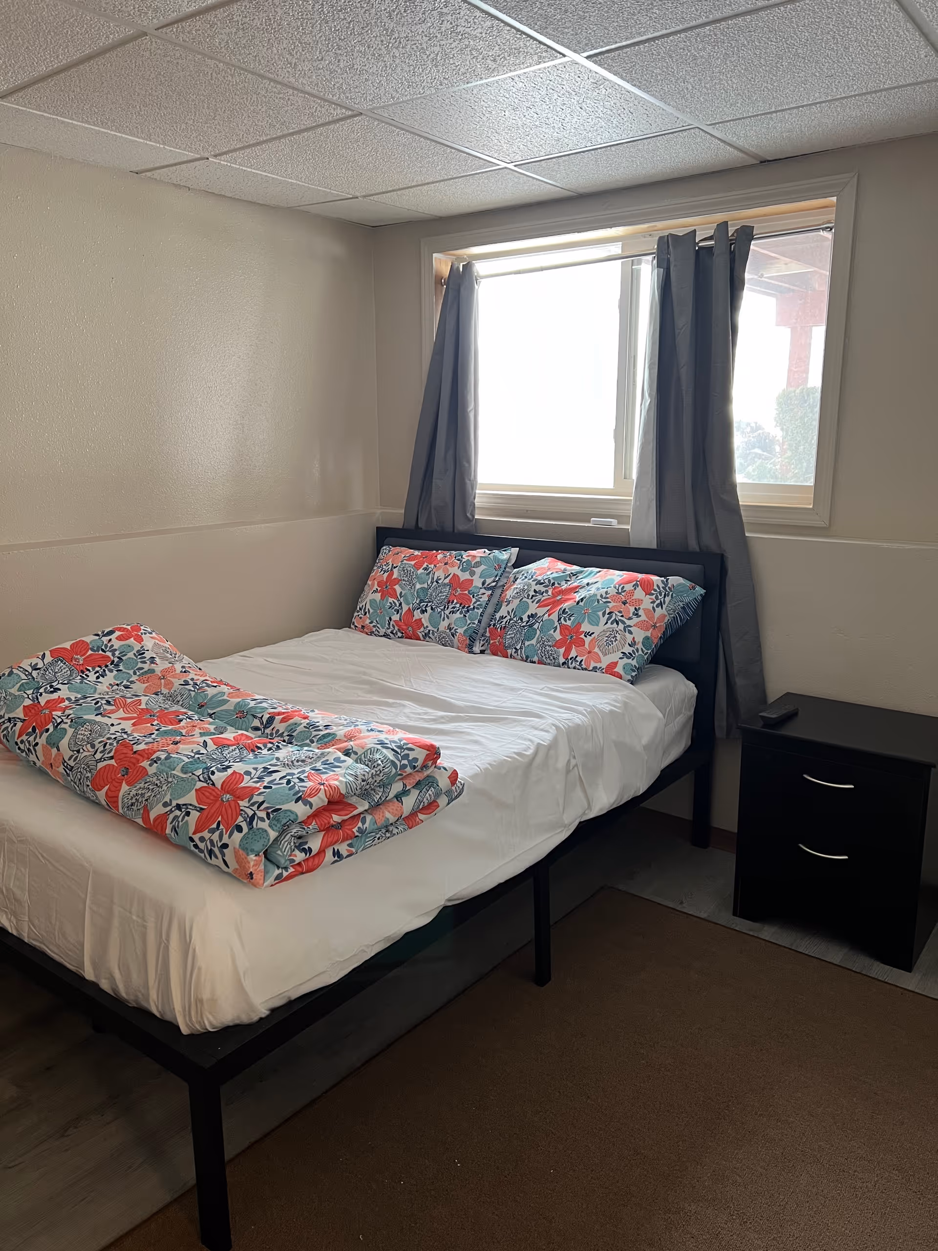 A small bedroom with a bed, floral bedding, window, and a black nightstand.