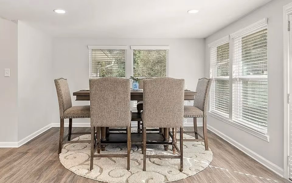 Bright dining room with a rectangular table and six upholstered chairs by windows.
