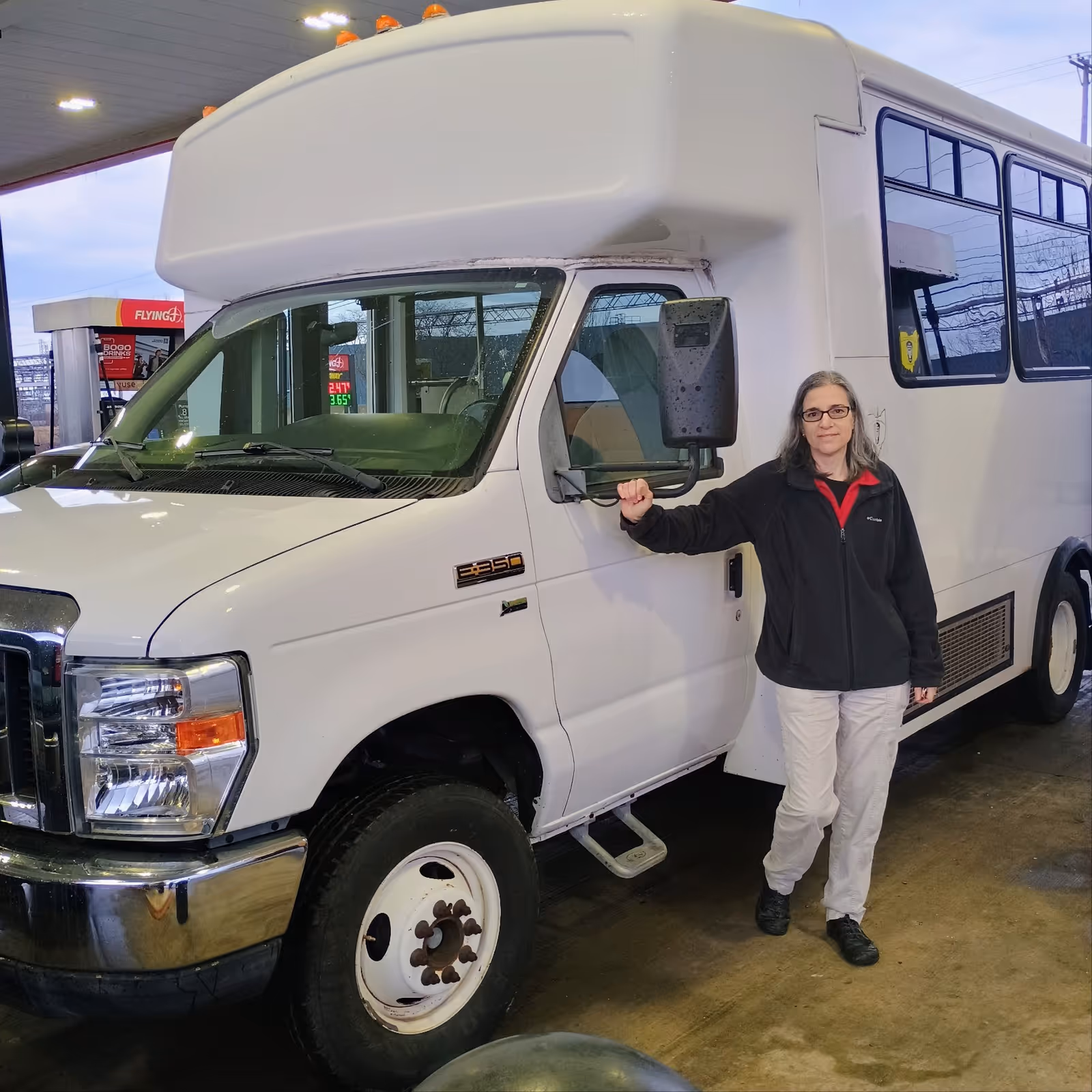 A woman stands beside a large white shuttle bus in a garage bay.