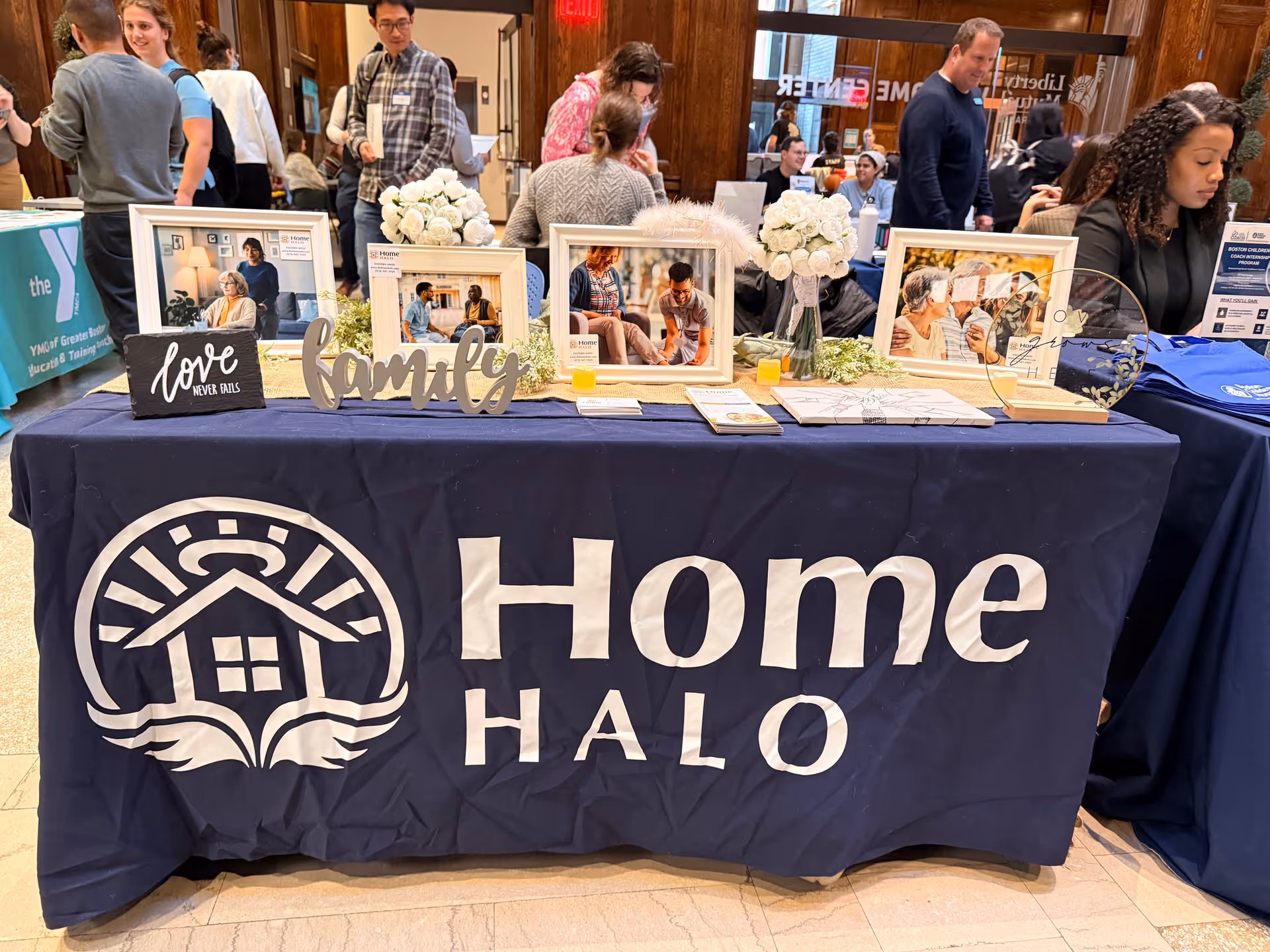 Indoor booth with Home Halo branding on a table, framed photos, and visitors.
