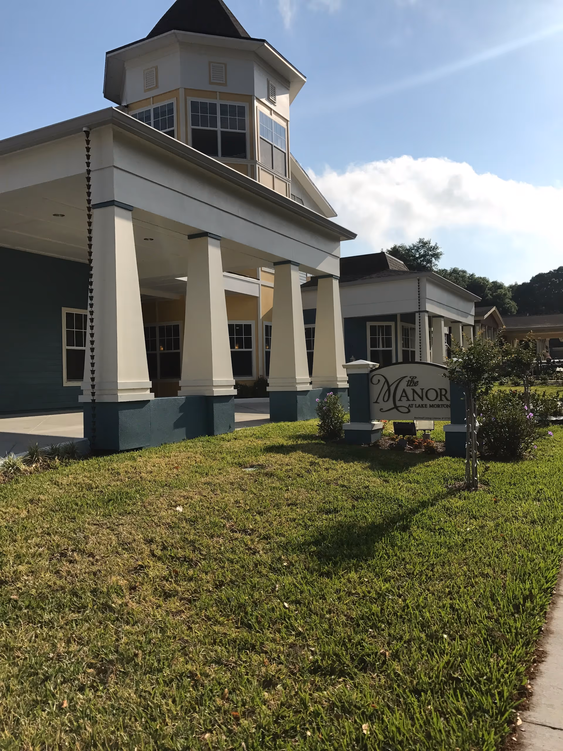 Exterior view of The Manor at Lake Morton building with a covered entrance supported by white pillars, a manicured lawn, and a sign displaying the facility's name. The sky is clear with some clouds and sunlight.