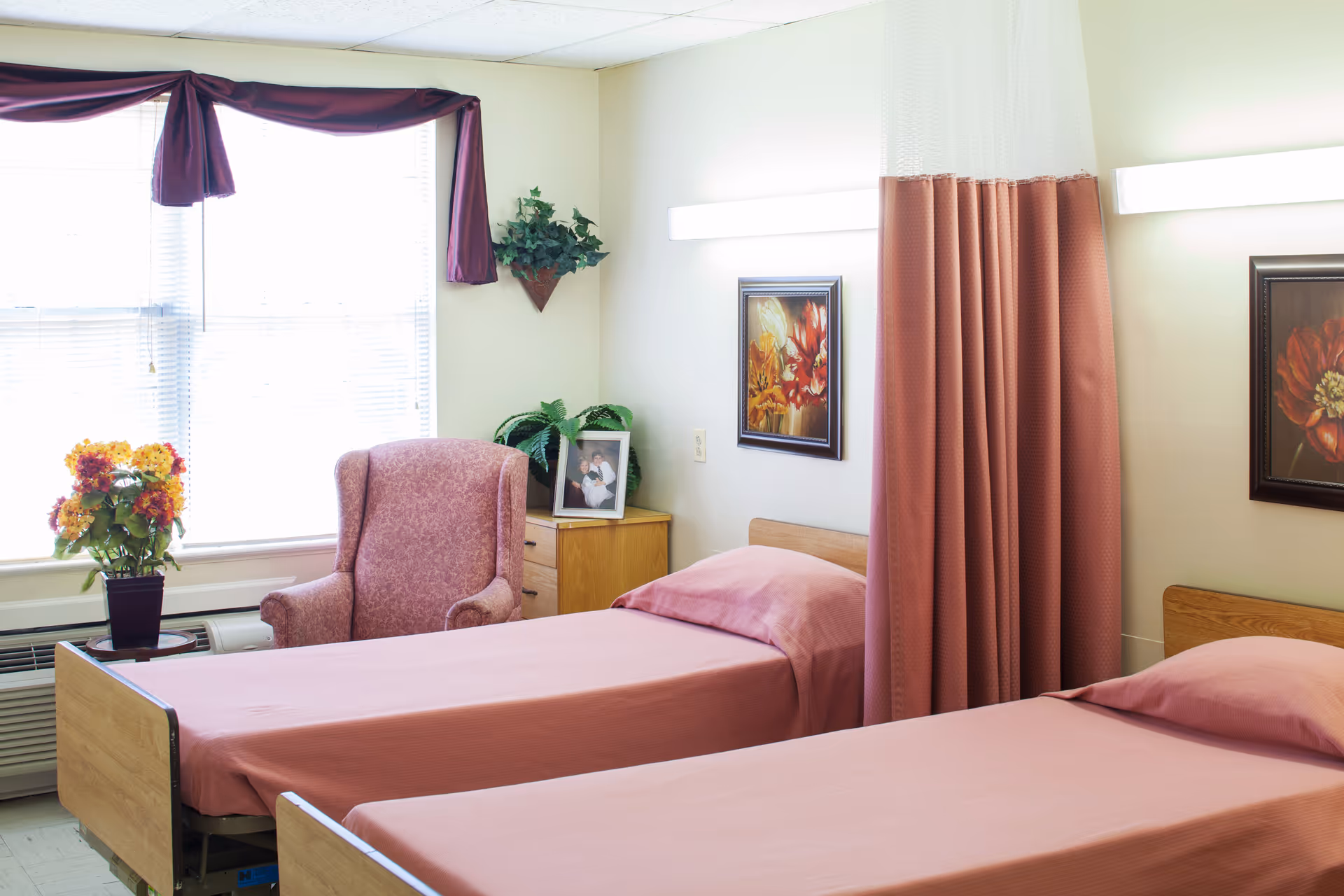 Two pink beds with a chair, flowers, and privacy curtains in a shared bedroom.