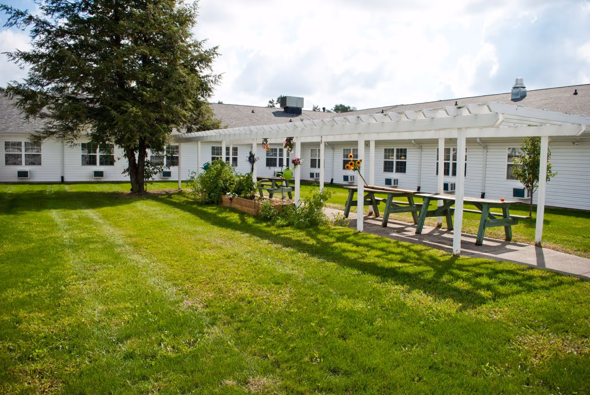 Outdoor garden area at Edgewater Woods featuring a green lawn, a large tree, a white pergola with hanging flower pots, picnic tables, and raised garden beds with plants and sunflowers, adjacent to a white building with multiple windows.