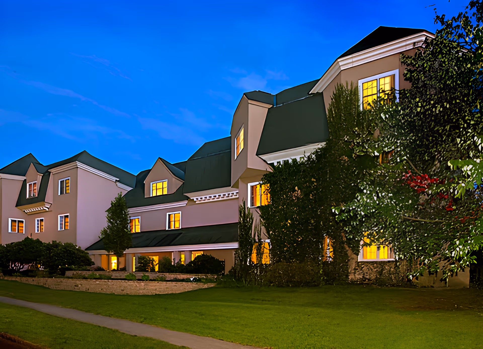 Front exterior of a multi-story senior living building at dusk with illuminated windows and landscaped lawn.