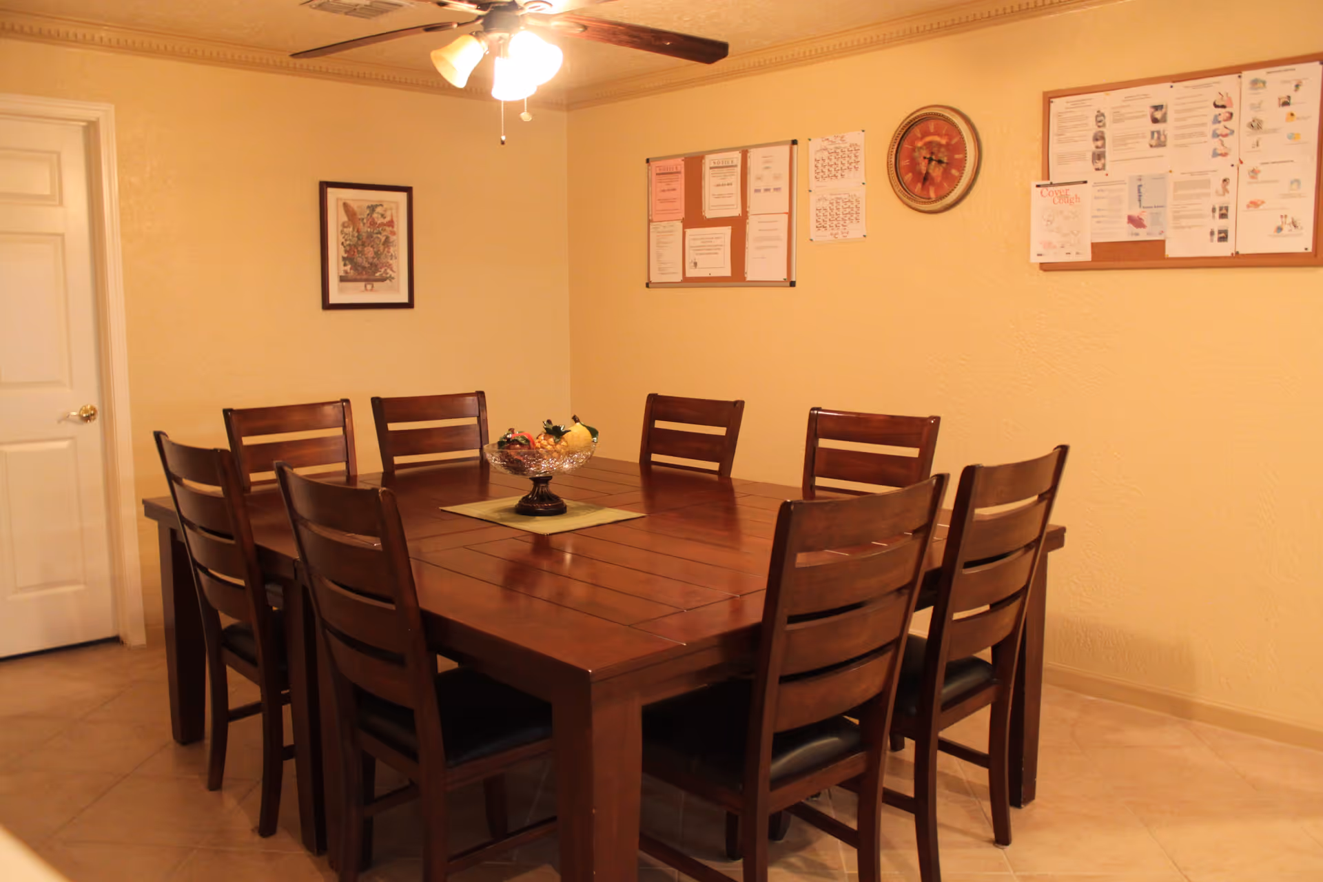 A dining room with a large wooden table surrounded by eight matching wooden chairs with black cushions. On the table is a decorative bowl with artificial fruit. The walls are painted beige and decorated with a framed picture, two bulletin boards with papers pinned on them, and a round wall clock. A ceiling fan with lights is mounted above the table. The floor is tiled and there is a closed white door in the background.