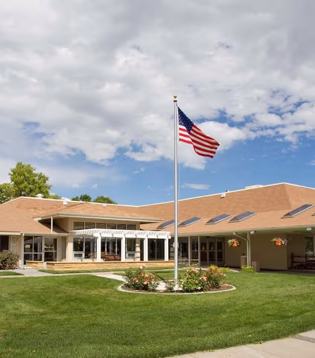Exterior view of a single-story nursing and rehabilitation facility with a brown roof, large windows, and a white pergola. In front of the building is a well-maintained lawn with a circular flower bed surrounding a tall flagpole flying the American flag. The sky is partly cloudy with patches of blue.
