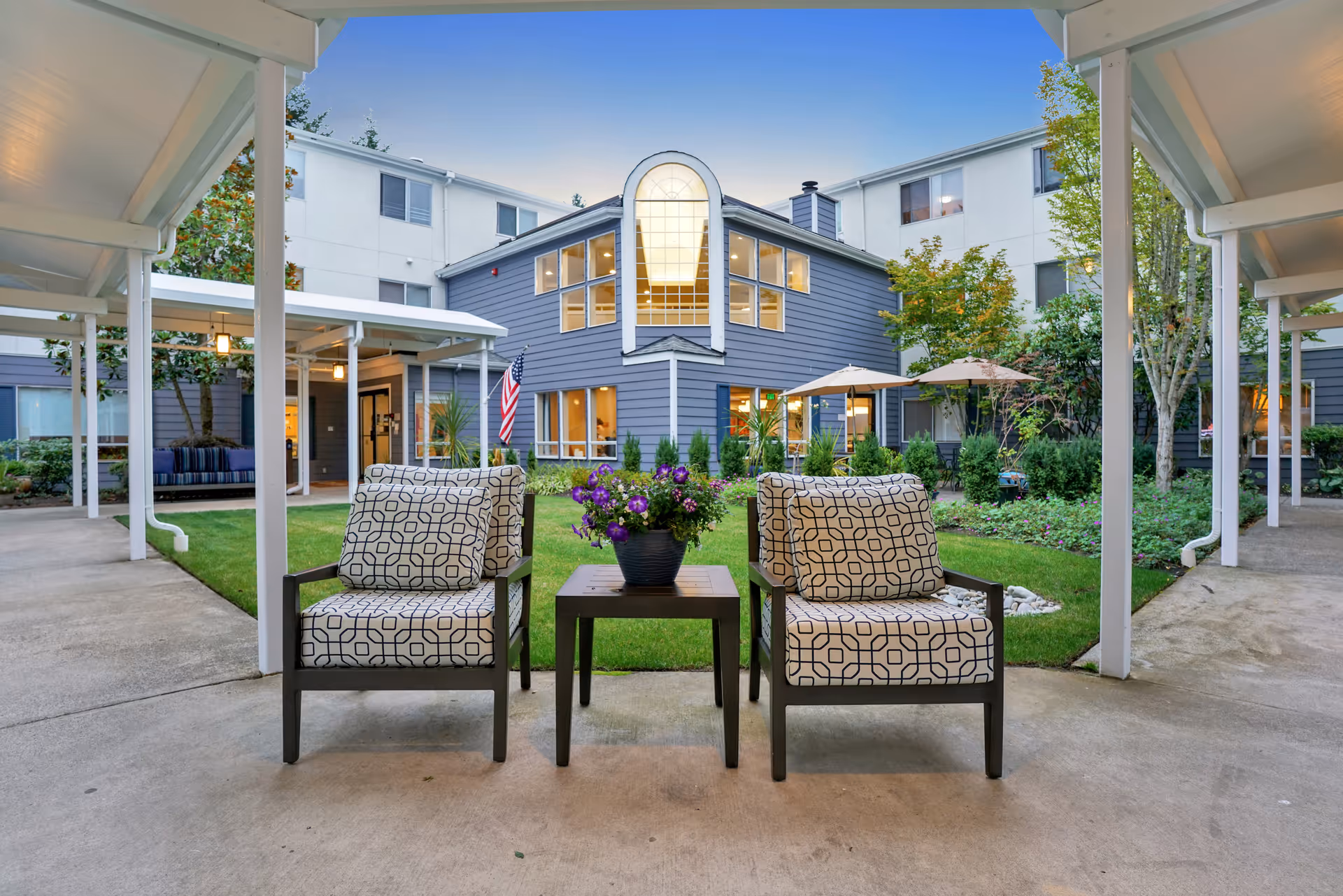 Outdoor seating area at Mountlake Terrace Plaza with two patterned cushioned chairs and a small table with a flower pot in the center, surrounded by a garden and a multi-story building in the background under a clear blue sky.