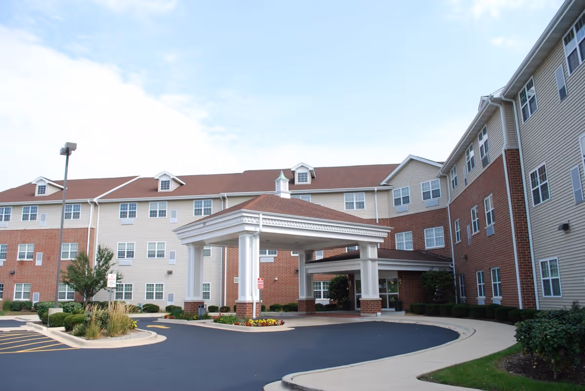Exterior view of a multi-story senior living facility building with beige siding and red brick accents. The building features many windows and a covered entrance with white columns and a red roof. There is a driveway and landscaped areas with bushes and flowers in front of the entrance.