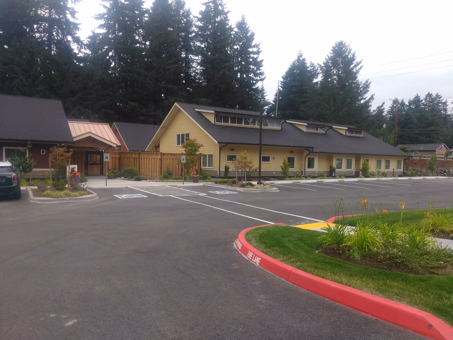 Single-story beige senior living building with a dark roof, large parking lot, and surrounding trees.