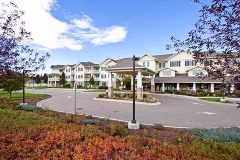 Front entrance and circular driveway of a multi-story beige retirement community building with landscaped grounds under a blue sky.