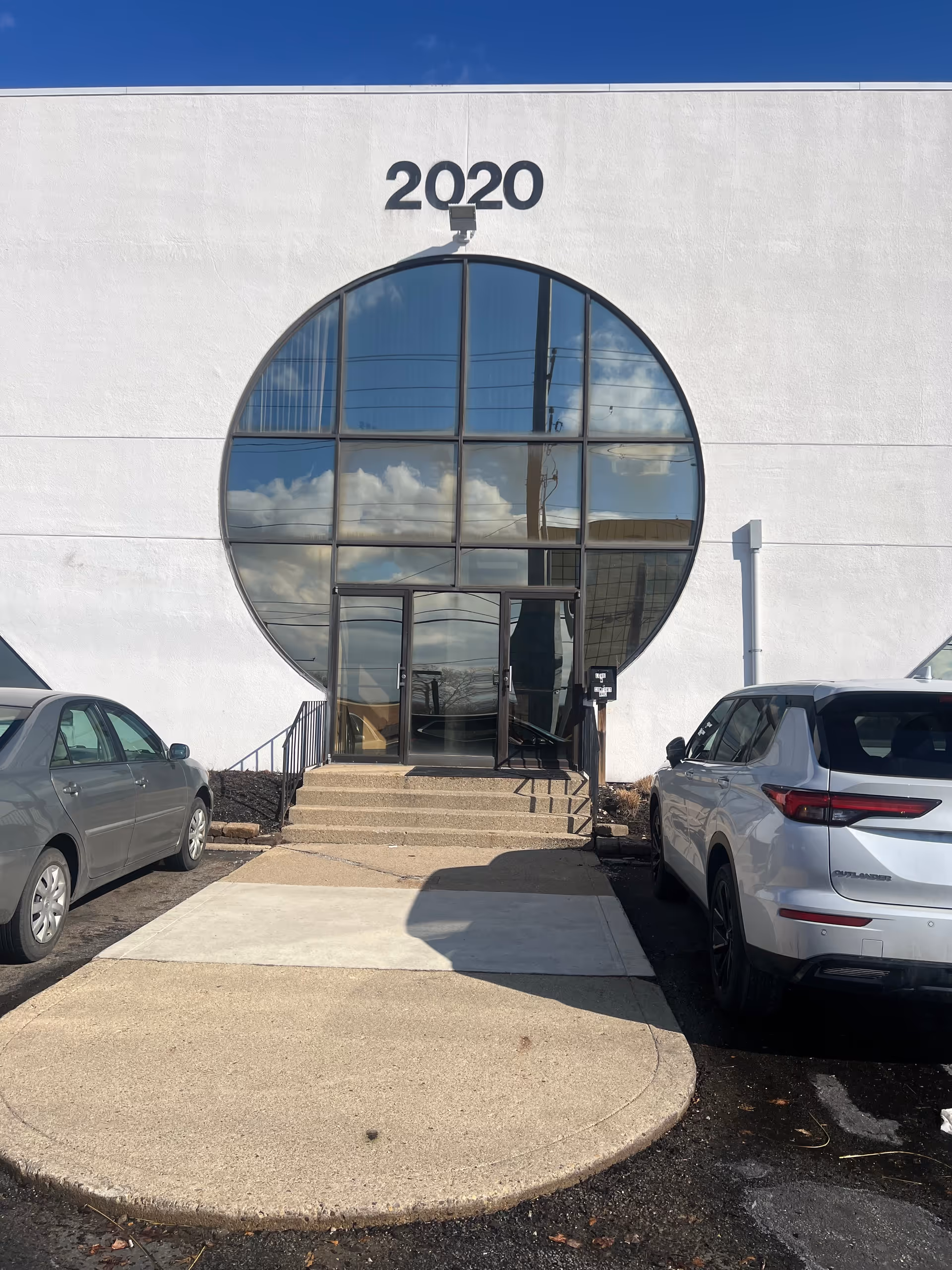 White building with a large circular glass entrance and 2020 above, parked cars nearby