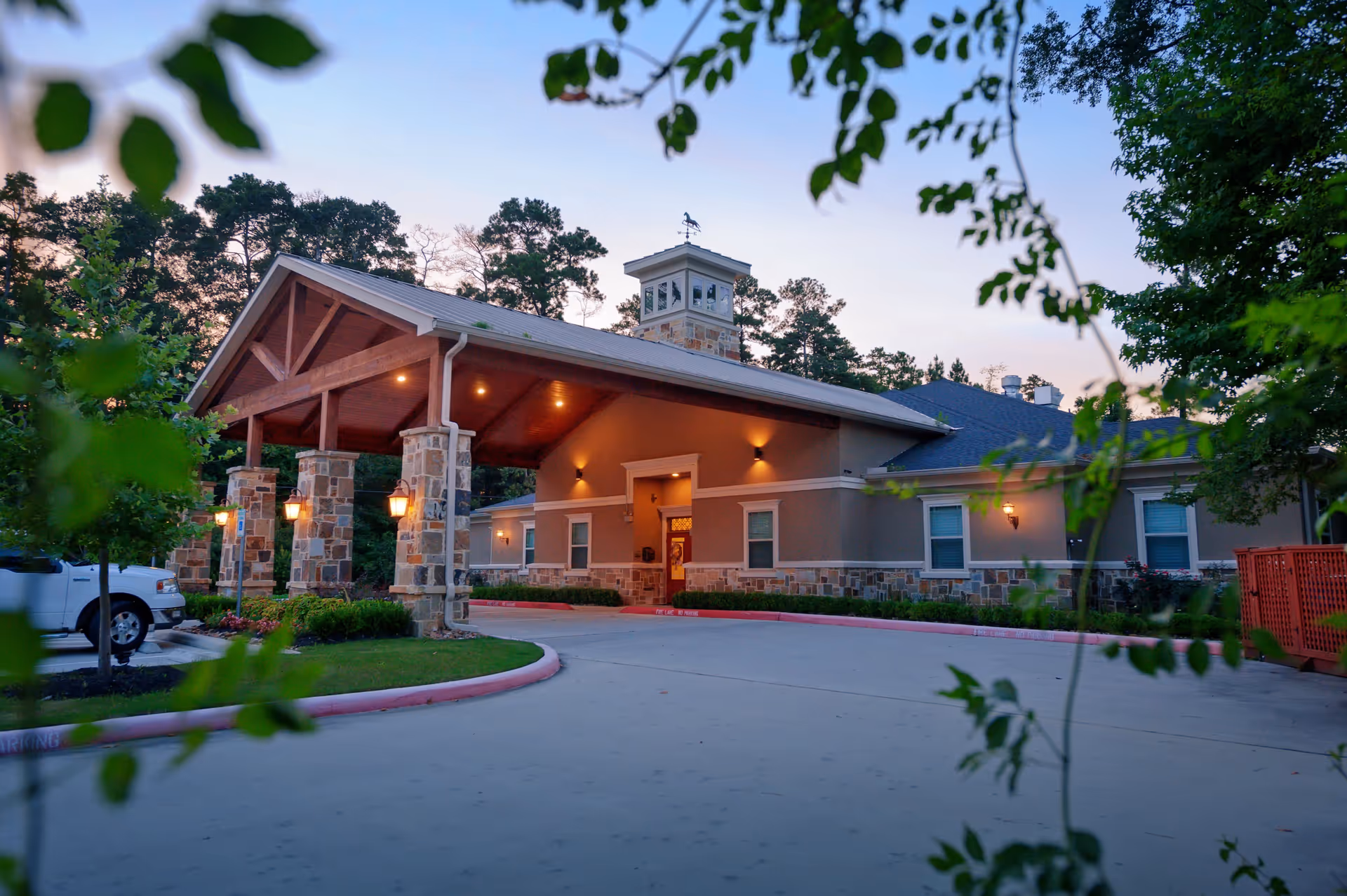 Front entrance of a single-story memory care building with a covered porte-cochere, stone pillars, and exterior lights framed by trees.