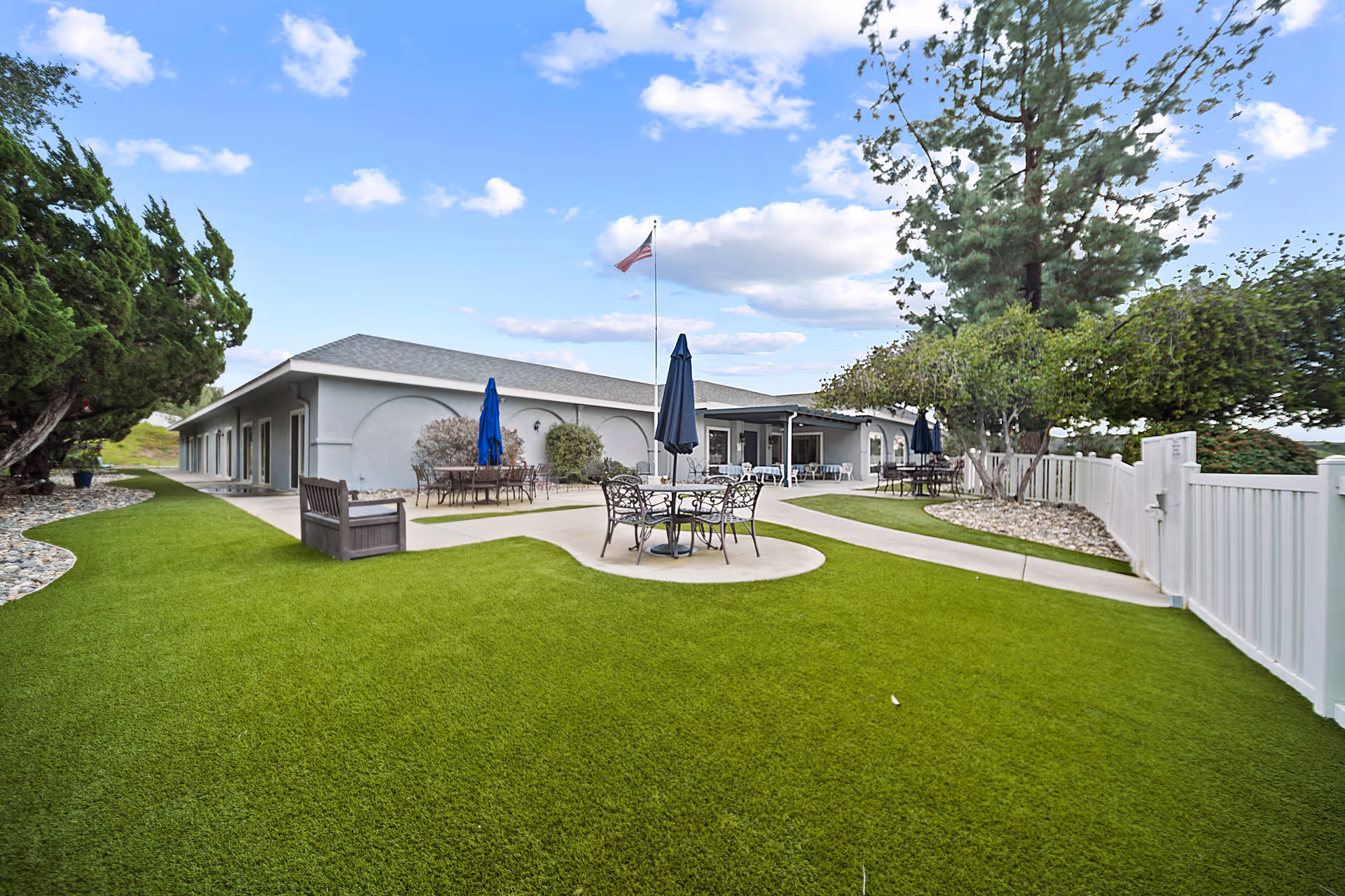 Outdoor patio area at Alpine View Lodge with green grass, several metal tables and chairs with closed umbrellas, a wooden bench, trees, and a white fence under a partly cloudy blue sky.