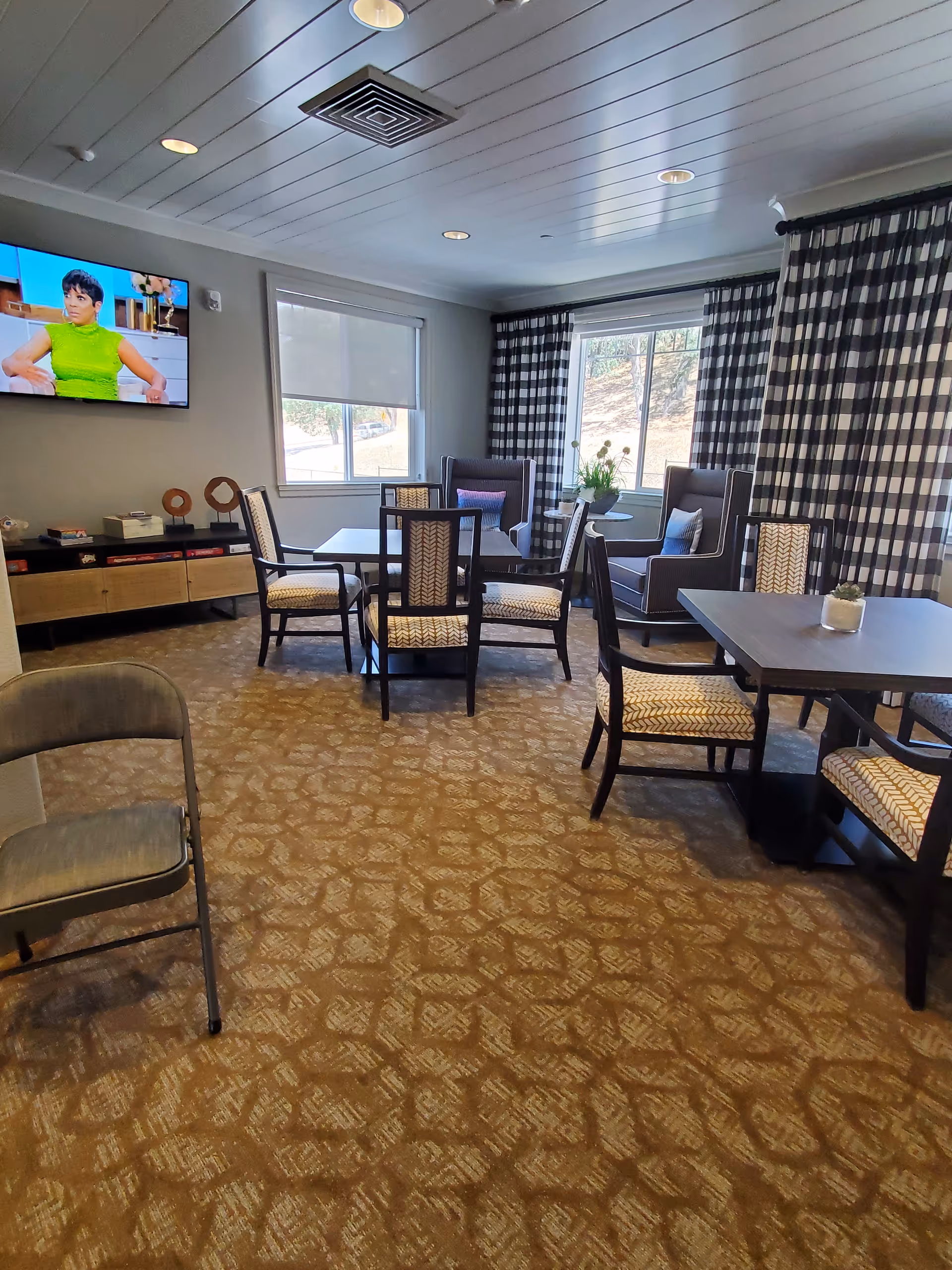 Senior living facility lounge with multiple tables, chairs, a TV, and large windows.