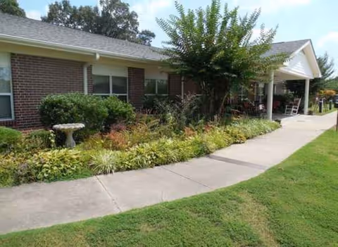 Exterior view of a single-story brick building with a covered porch area, surrounded by a well-maintained garden with various plants and shrubs, and a concrete walkway leading up to the entrance.