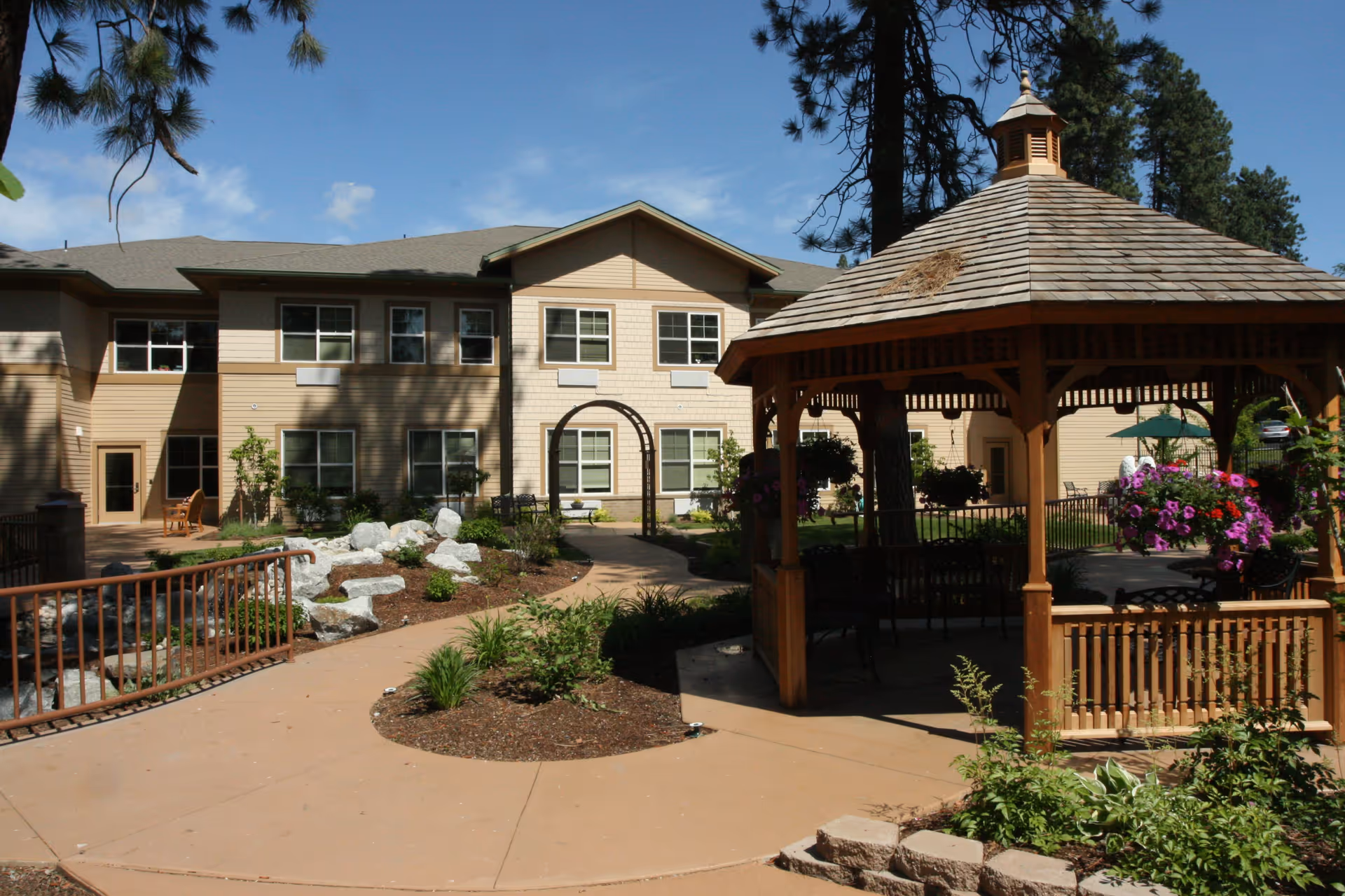 Outdoor courtyard area of Orchard Ridge Senior Living featuring a wooden gazebo with hanging flower baskets, a paved walkway, landscaped garden beds with rocks and plants, and a two-story beige building in the background under a clear blue sky.