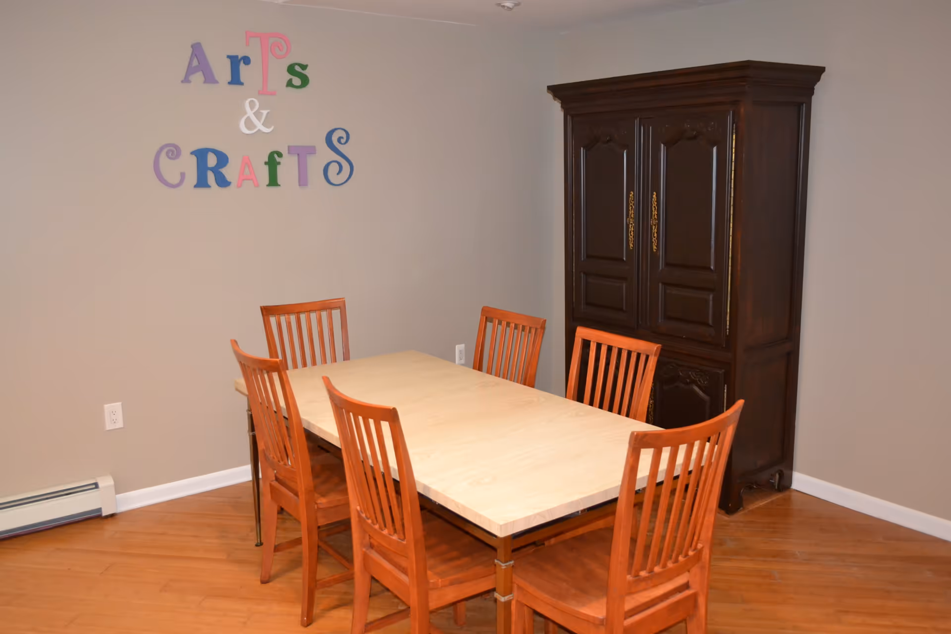 A room with a rectangular table surrounded by six wooden chairs. On the wall above the table, colorful letters spell out 'Arts & Crafts'. There is a large dark wooden cabinet against the wall to the right. The floor is wooden and the walls are painted beige.