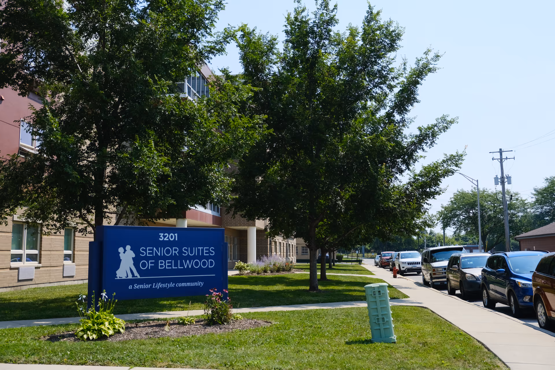 Front exterior of the Senior Suites of Bellwood building with a blue entrance sign, trees, and cars parked along the sidewalk.