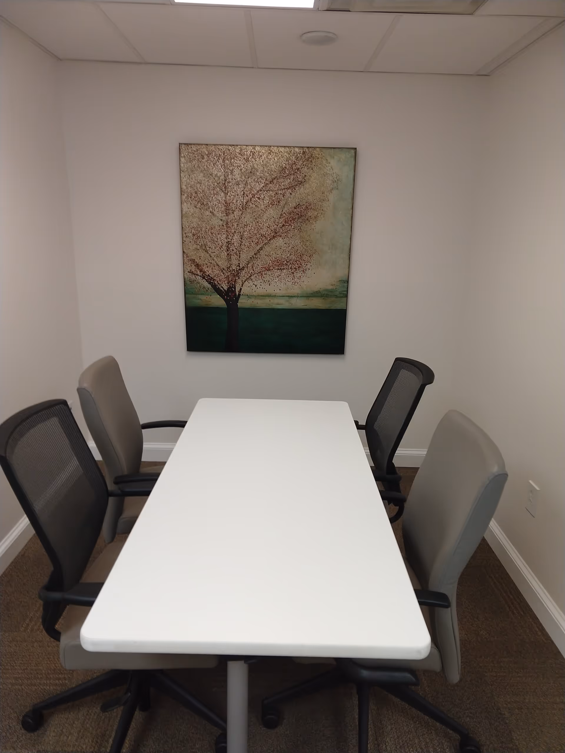 Small meeting room with a white table, six office chairs, and a tree artwork on wall.